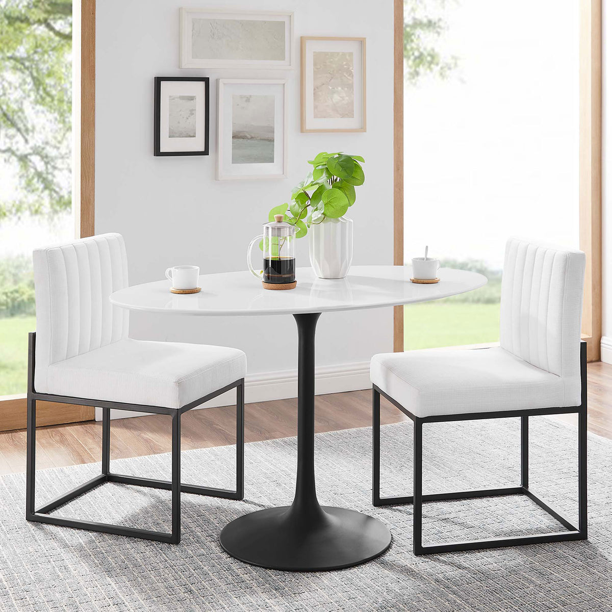 Compact white oval dining table with black pedestal base styled with two modern chairs in a bright room || EEI-3517-BLK-WHI || front_angle_room