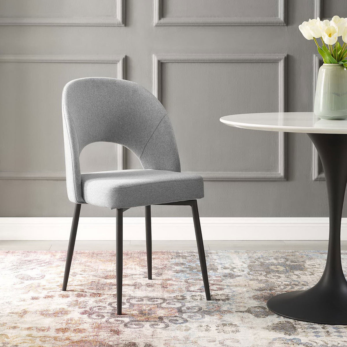 Gray upholstered dining chair with cutout back arranged beside a round pedestal dining table and floral decor. || EEI-3801-BLK-LGR || front_angle_room