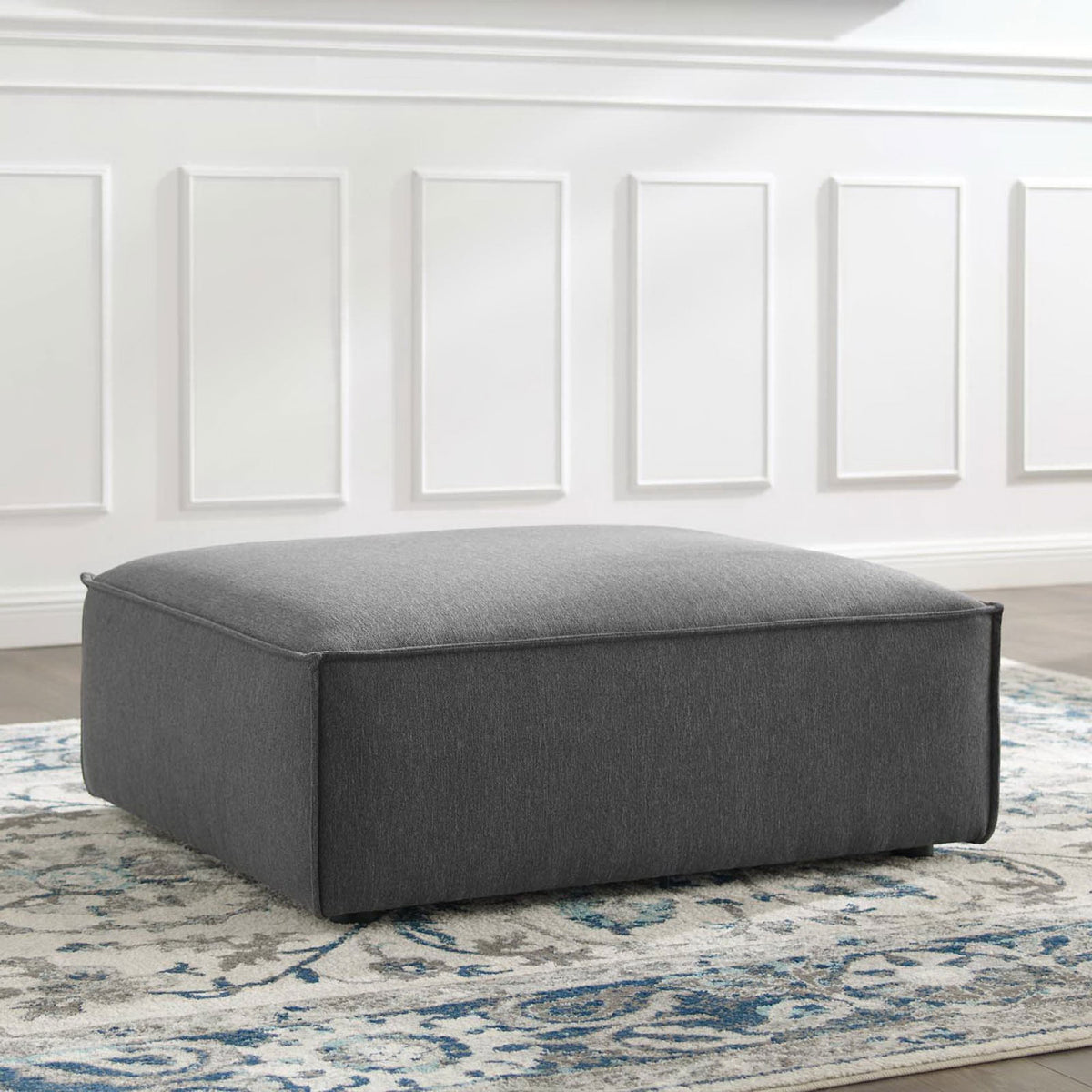 Charcoal upholstered ottoman styled on a patterned rug to add modern comfort to a bright living space || EEI-3873-CHA || front_angle_room