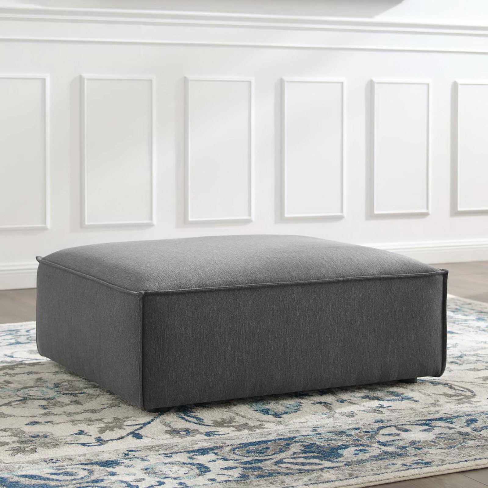 Charcoal upholstered ottoman styled on a patterned rug to add modern comfort to a bright living space || EEI-3873-CHA || front_angle_room