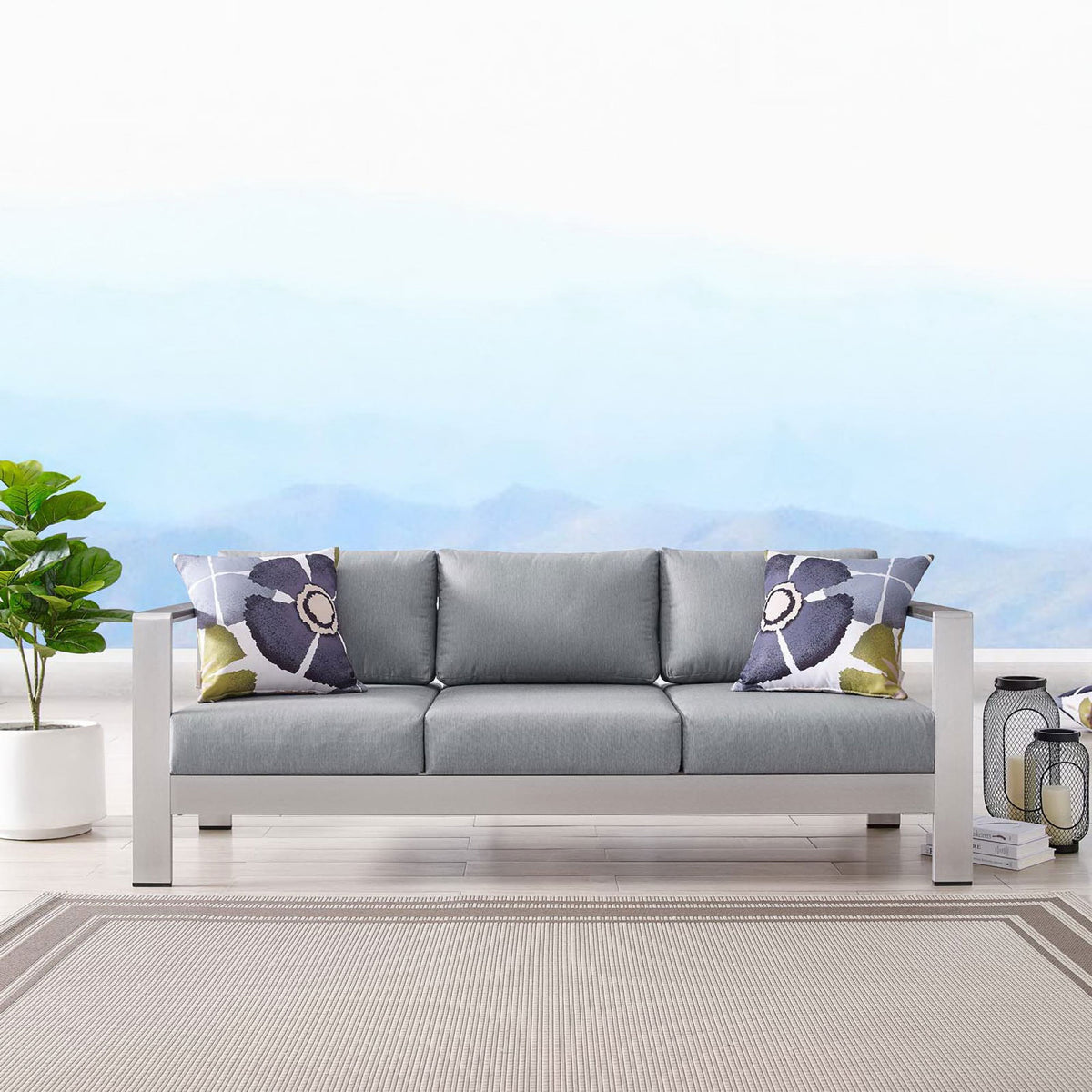 Gray cushioned sofa styled with patterned pillows, lanterns, and a neutral outdoor rug || EEI-3917-SLV-GRY || front_room