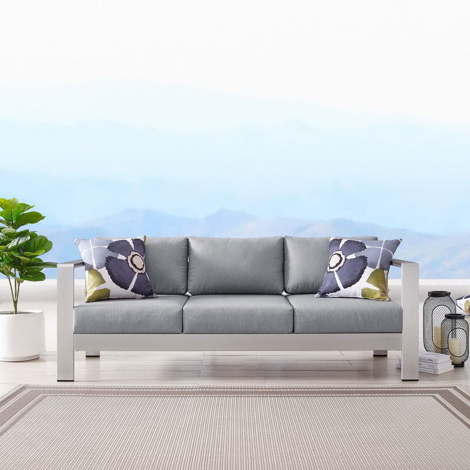 Gray cushioned sofa styled with patterned pillows, lanterns, and a neutral outdoor rug || EEI-3917-SLV-GRY || front_room