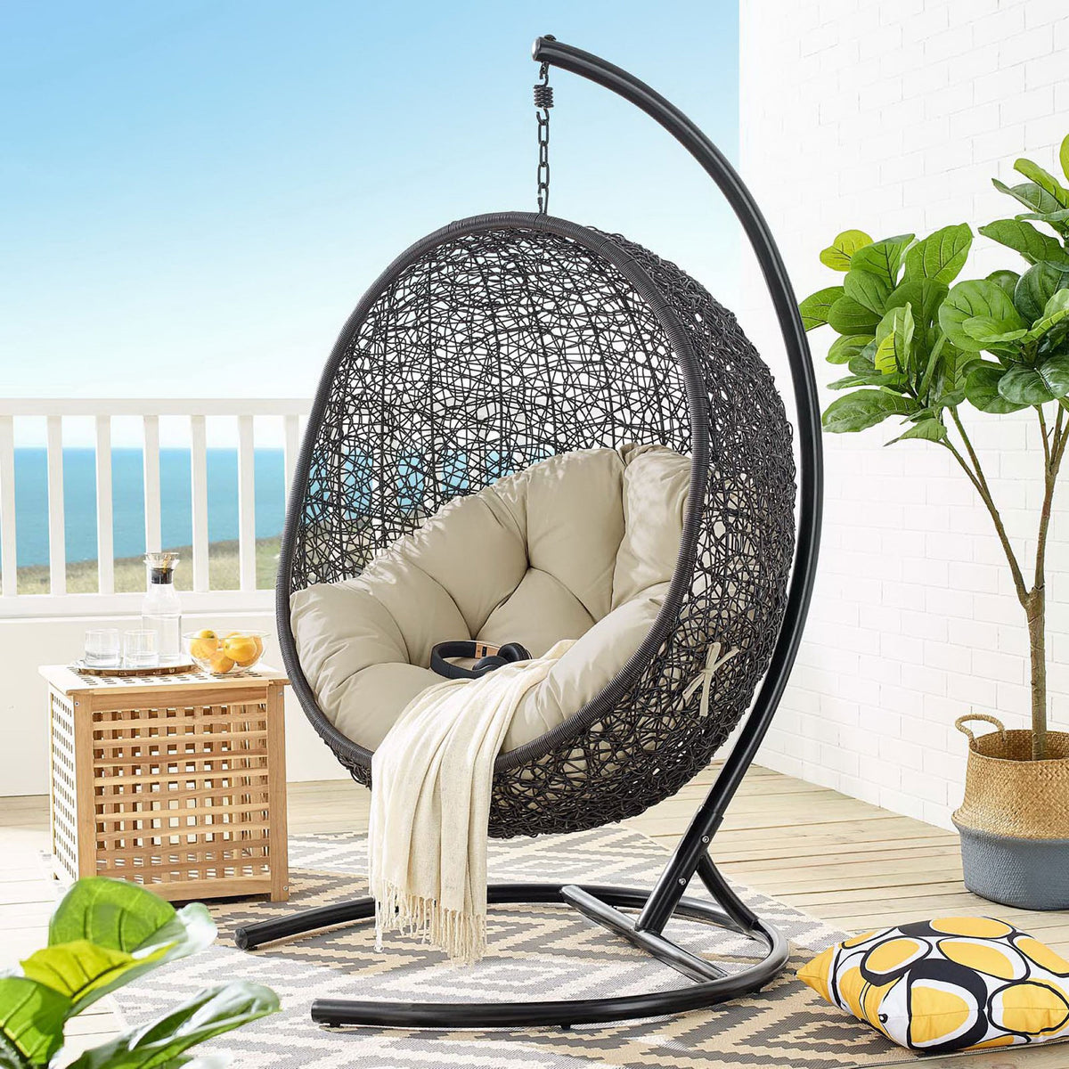 Beige cushioned hanging egg chair with stand styled on a coastal patio with rug and greenery || EEI-3943-BLK-BEI || front_angle_room