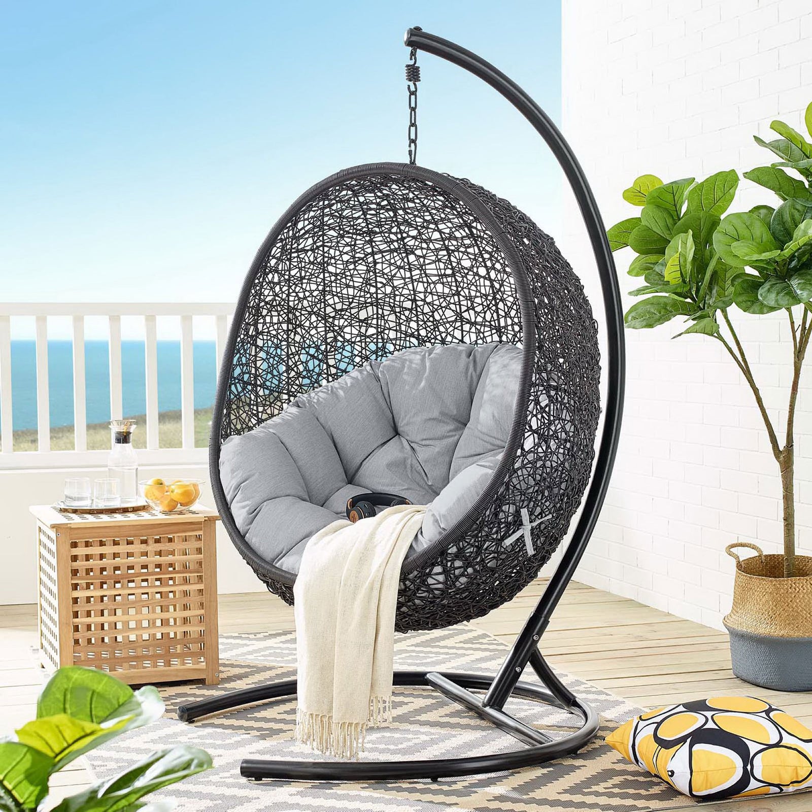 Gray cushioned hanging egg chair and stand styled on a sunny balcony with coastal decor || EEI-3943-BLK-GRY || front_angle_room