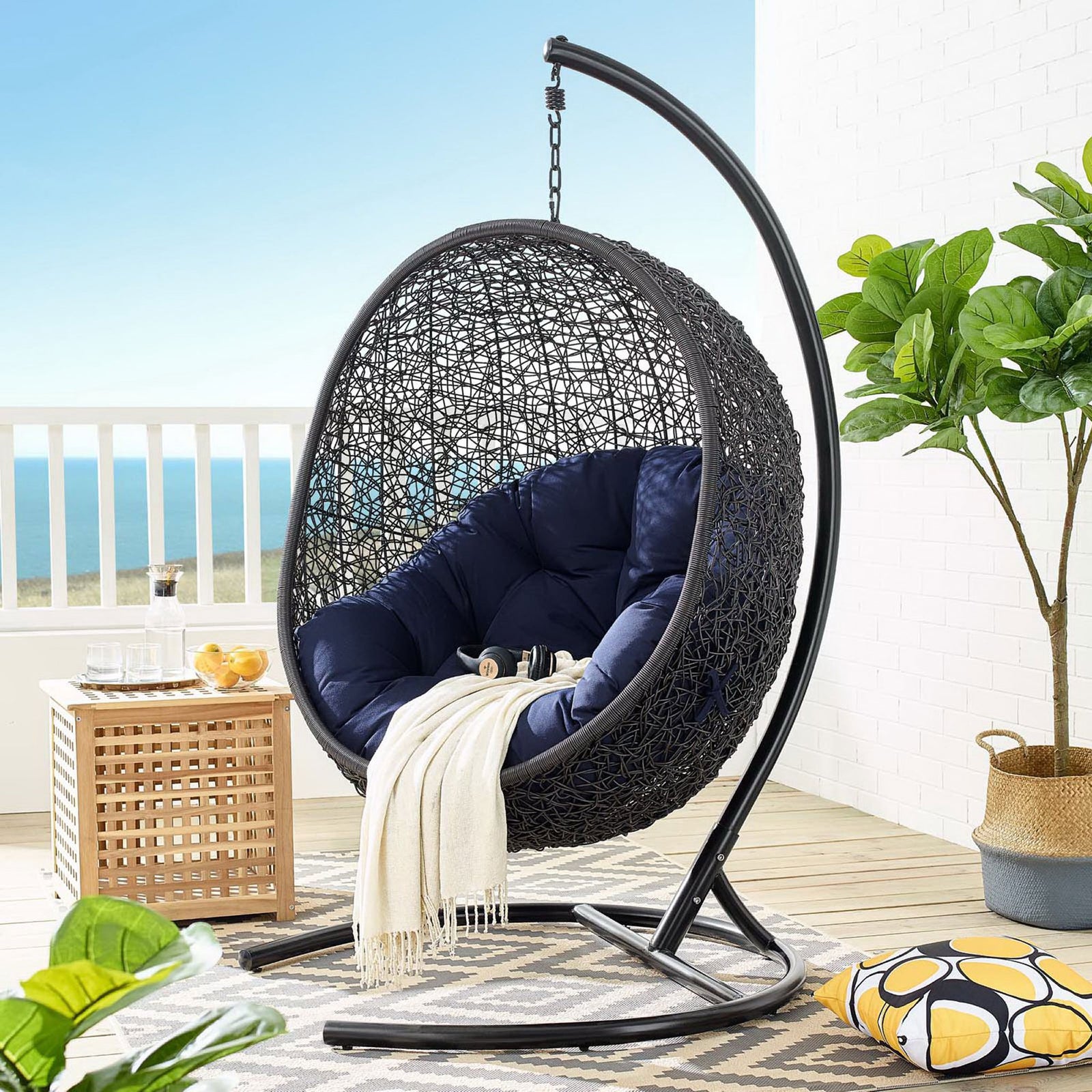 Navy cushioned hanging egg chair and stand styled on a breezy coastal balcony with rug || EEI-3943-BLK-NAV || front_angle_room