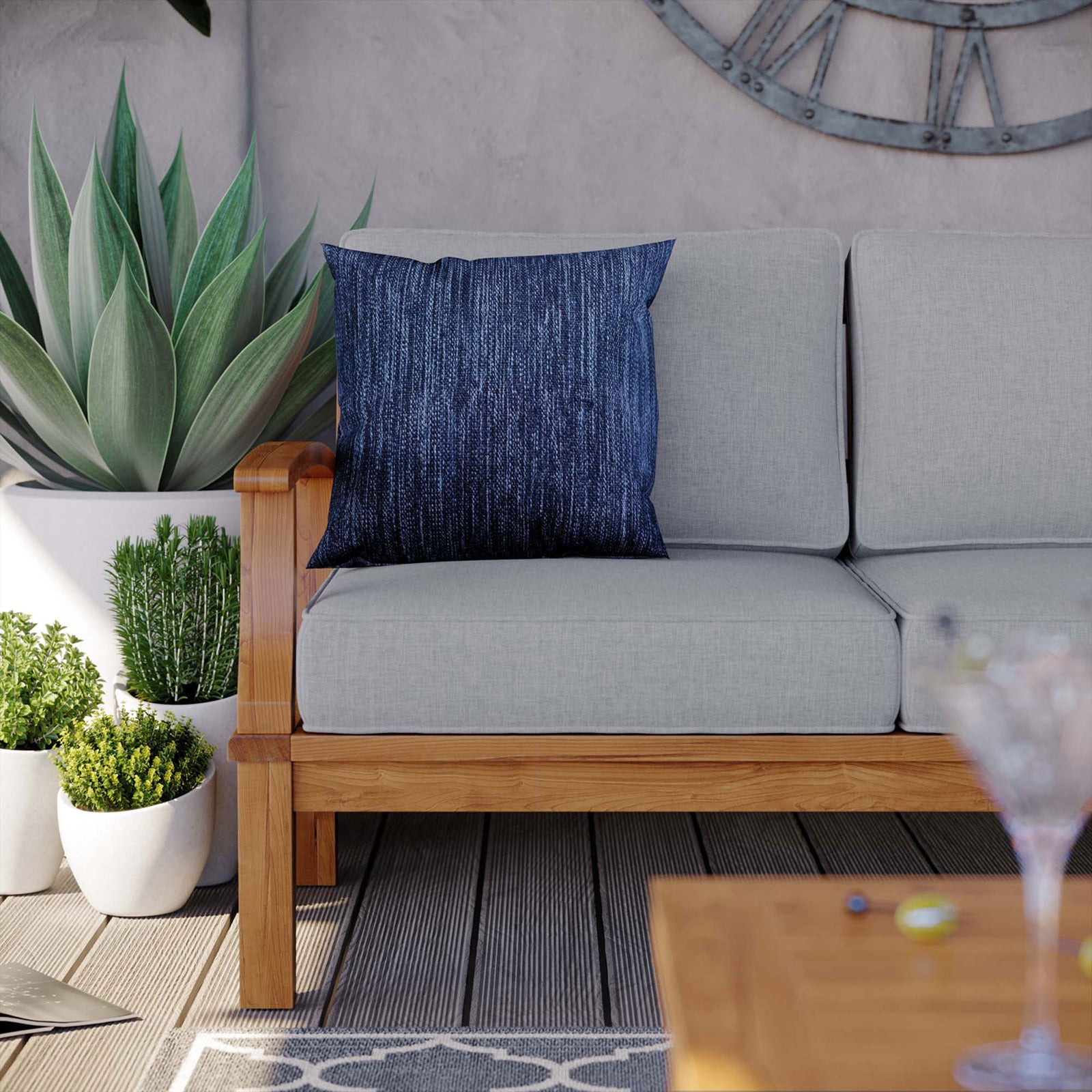 Teak patio sofa corner with gray cushions, navy accent pillow, and lush potted greenery || EEI-4176-NAT-GRY || detail_room