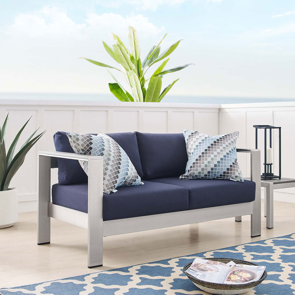 Navy cushioned aluminum loveseat styled with patterned pillows in a bright coastal-inspired room || EEI-4226-SLV-NAV || front_angle_room
