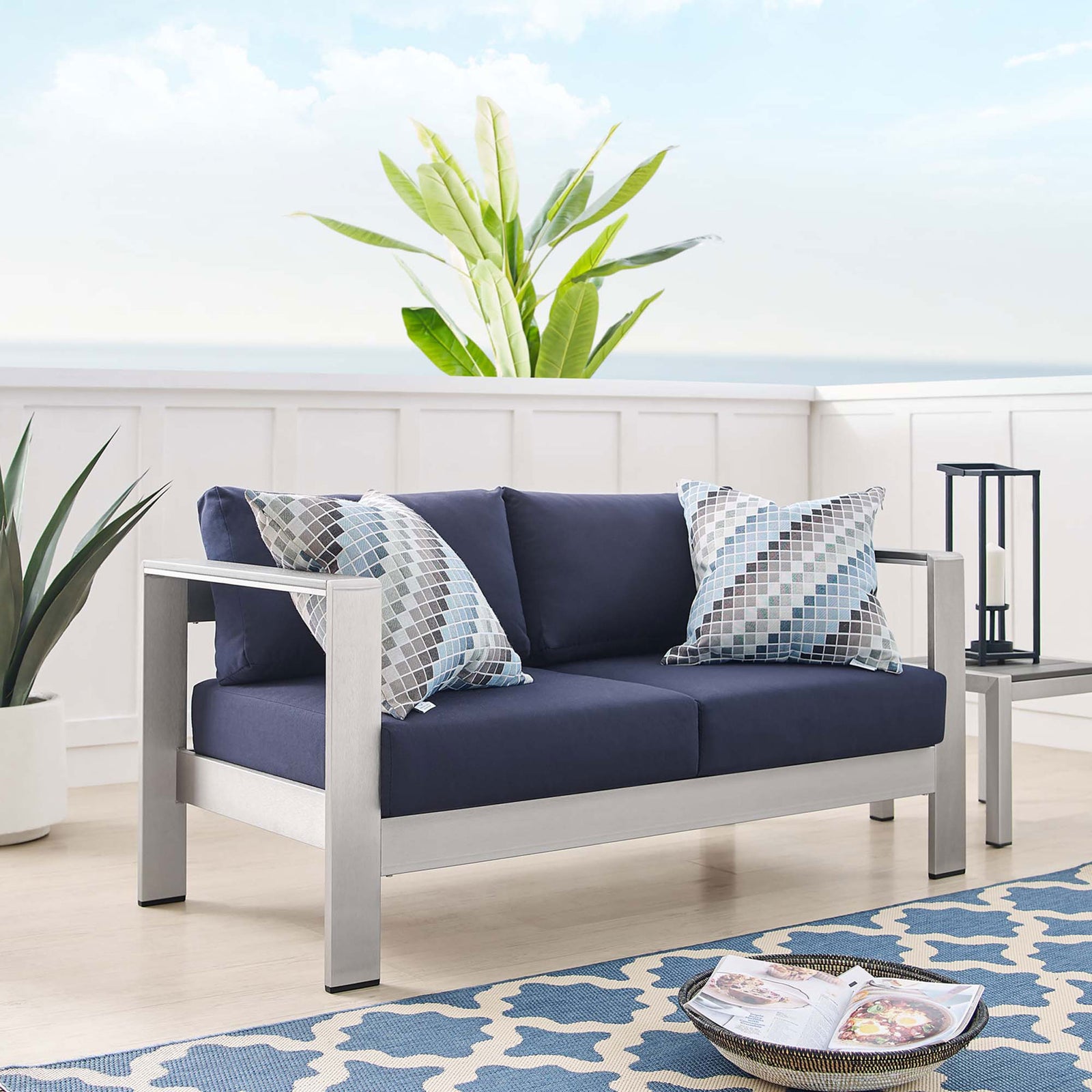 Navy cushioned aluminum loveseat styled with patterned pillows in a bright coastal-inspired room || EEI-4226-SLV-NAV || front_angle_room