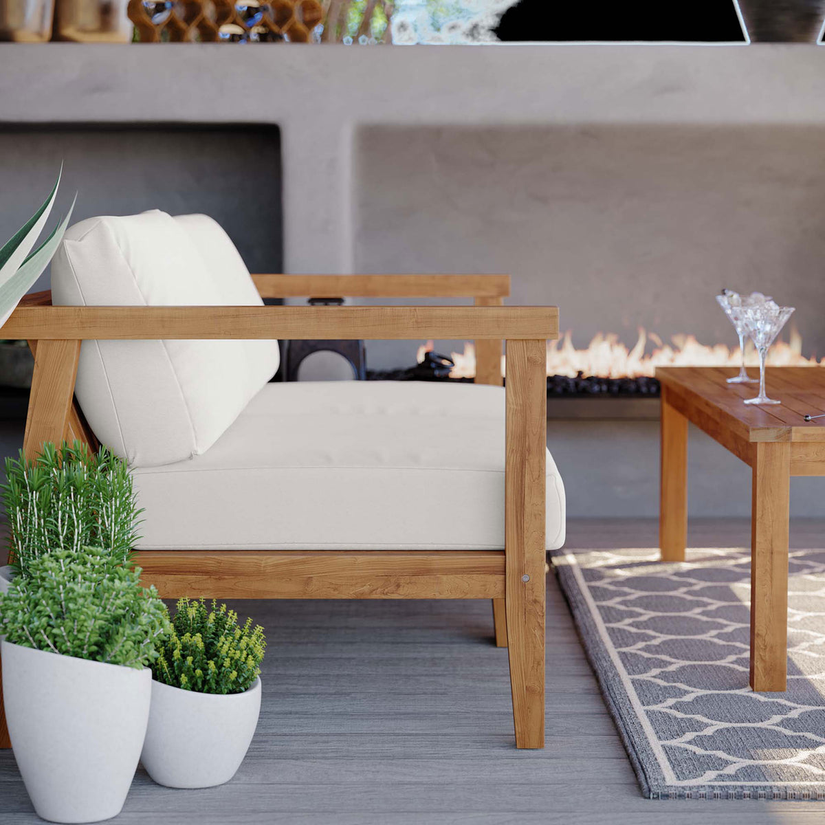 Bayport teak chair set by a modern outdoor fireplace with white cushions and lush green plants || EEI-4259-NAT-WHI-SET || front_angle_partial_room