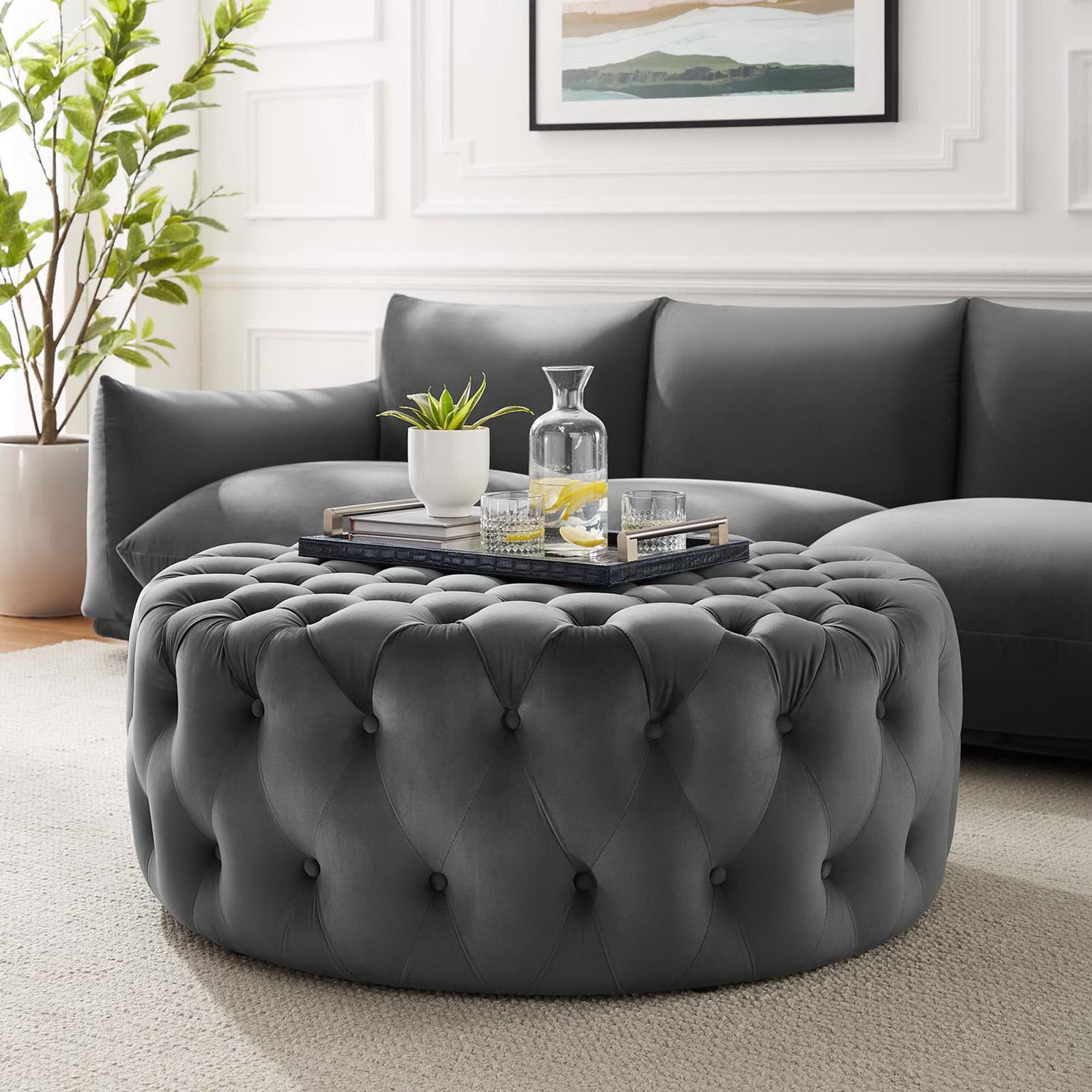 Large round gray velvet ottoman styled in a bright living room as a plush centerpiece and versatile coffee table || EEI-5469-GRY || front_angle_room