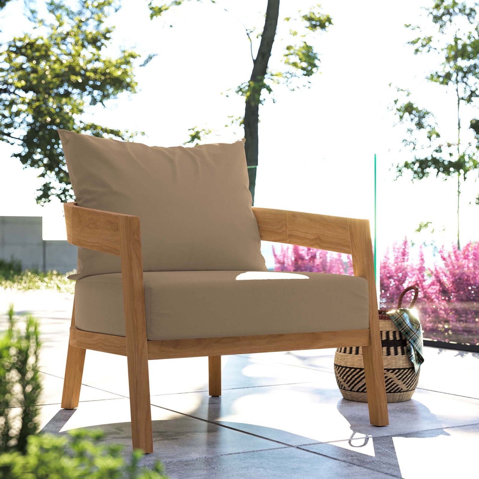 Light brown cushioned teak patio armchair on a sunlit terrace framed by greenery and blooming flowers || EEI-5602-NAT-LBR || front_angle_room
