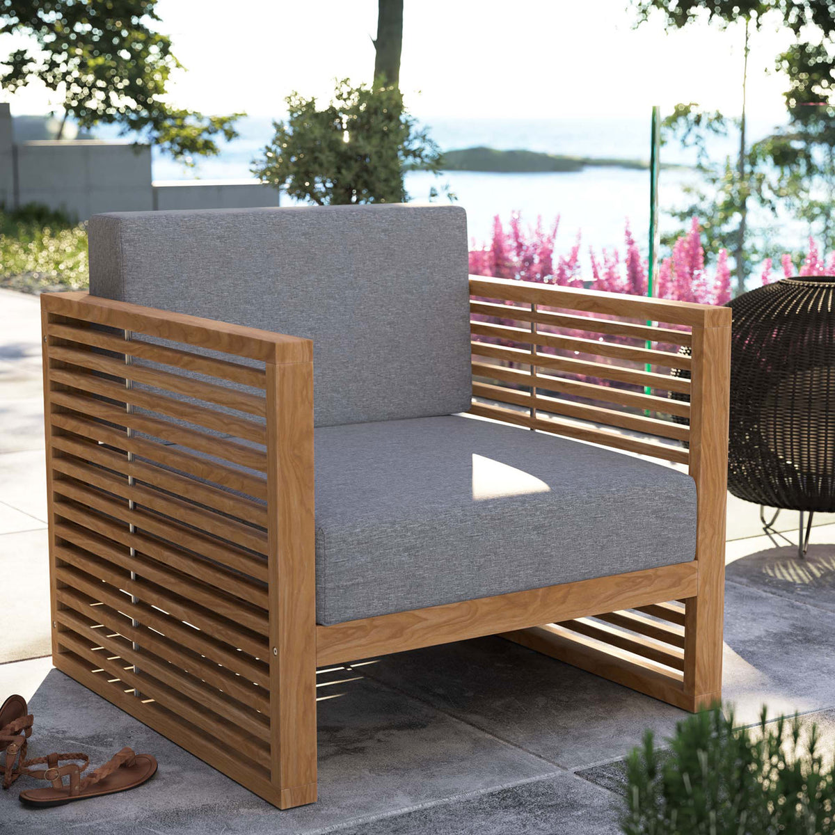 Carlsbad teak outdoor armchair with gray cushions styled on a sunny stone patio overlooking water. || EEI-5606-NAT-GRY || front_angle_room