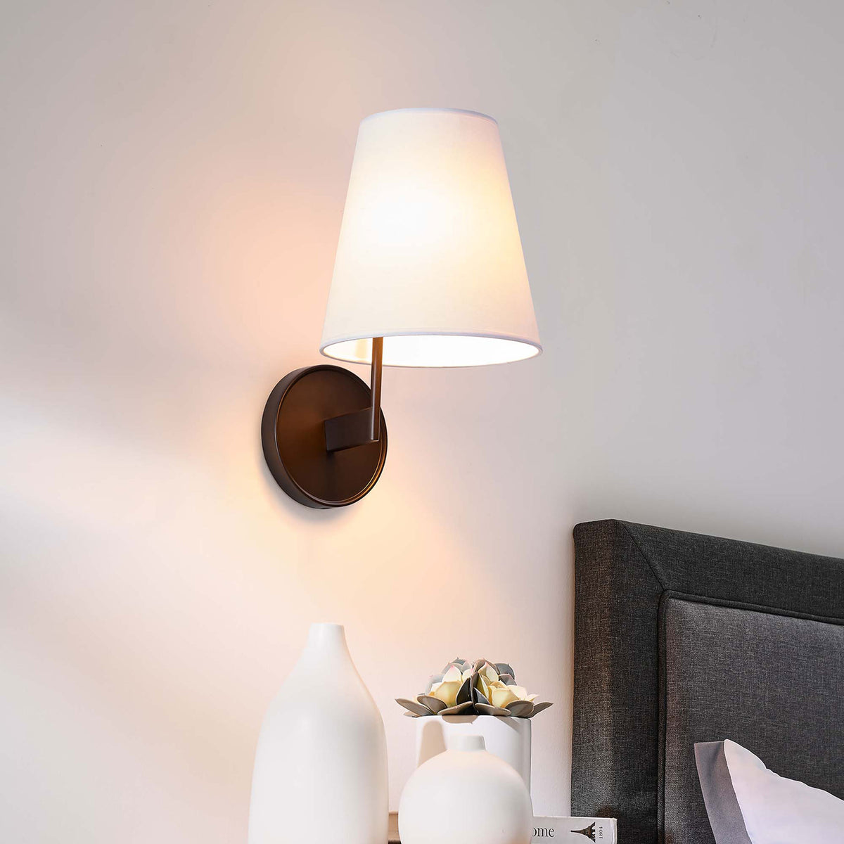 Bronze wall sconce beside a bed providing soft ambient light over a nightstand vignette. || EEI-5643-WHI-BRZ || front_angle_on_room