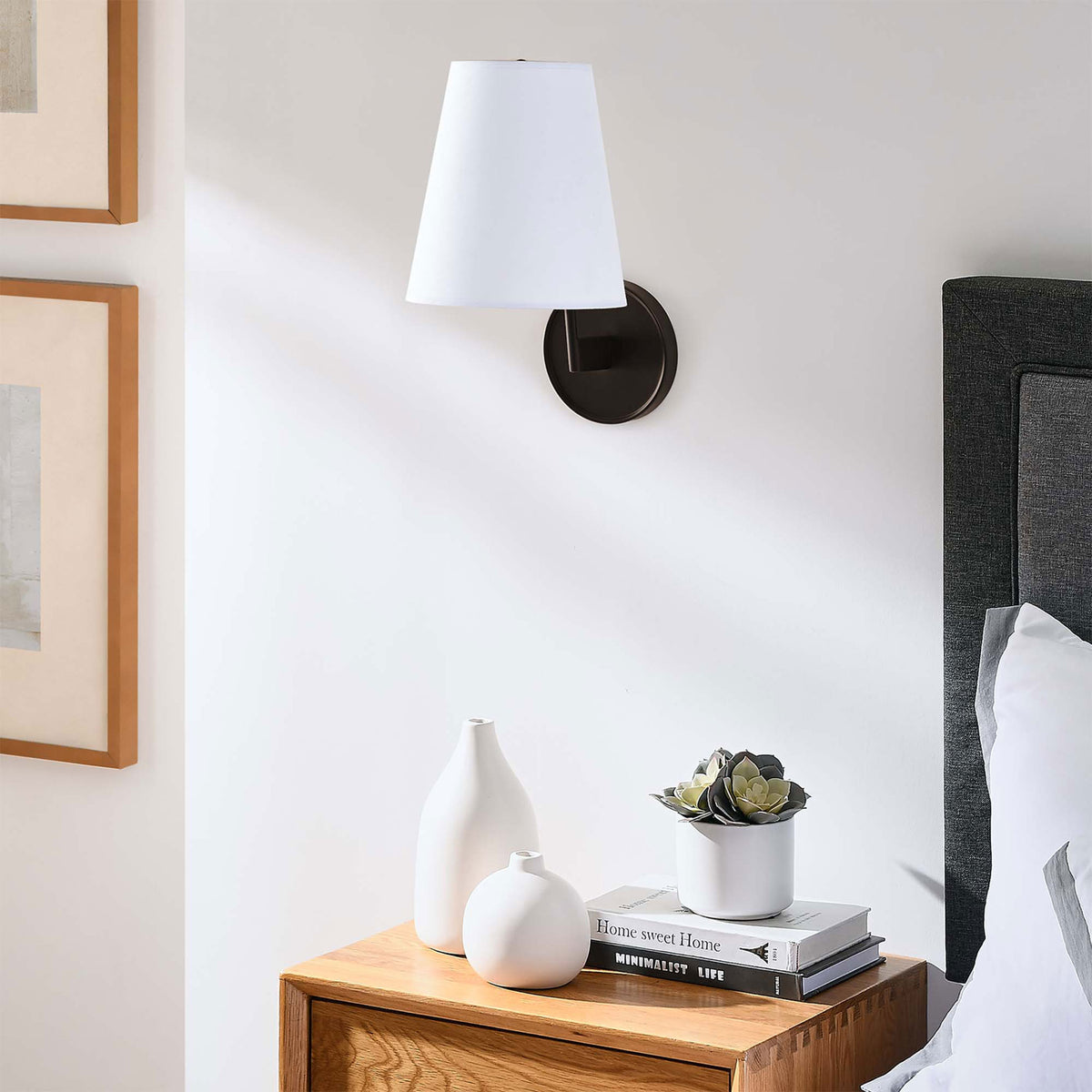 Bronze wall sconce with white shade mounted above a nightstand for modern bedroom decor. || EEI-5643-WHI-BRZ || front_angle_room