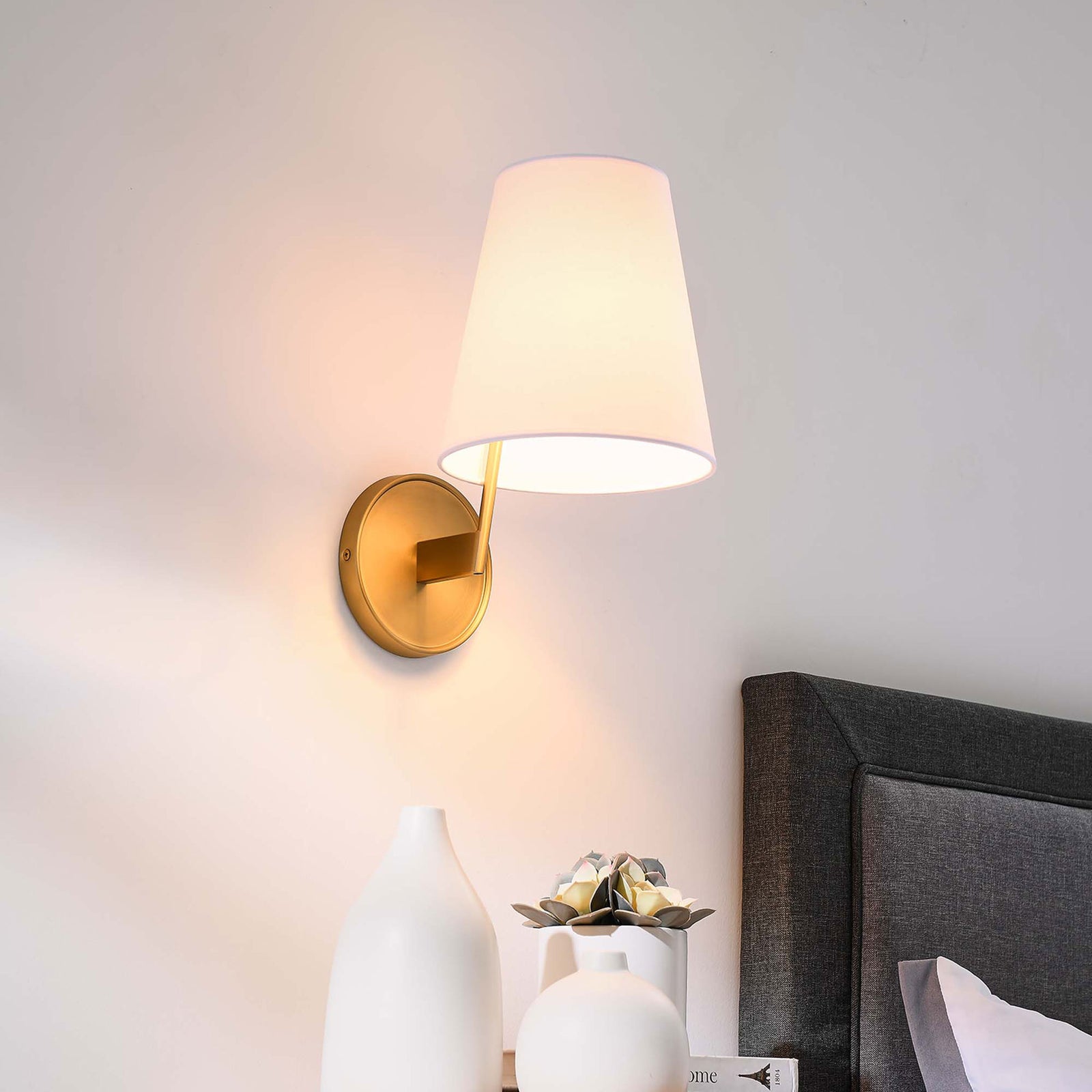 Satin brass wall sconce beside a bed illuminating a nightstand arrangement with warm light. || EEI-5643-WHI-SBR || front_angle_on_room