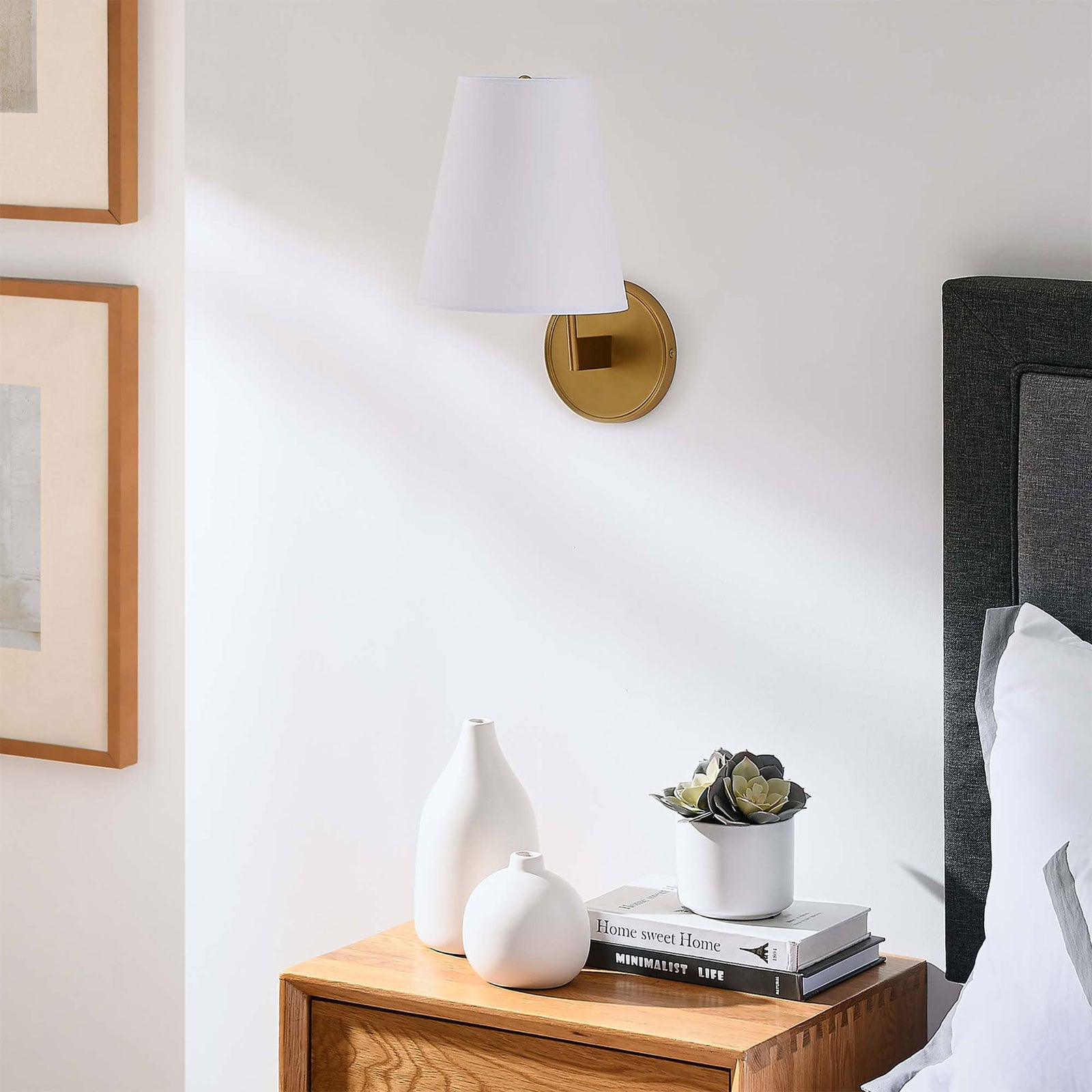 Satin brass wall sconce with white shade mounted above a nightstand in a bright bedroom. || EEI-5643-WHI-SBR || front_angle_room