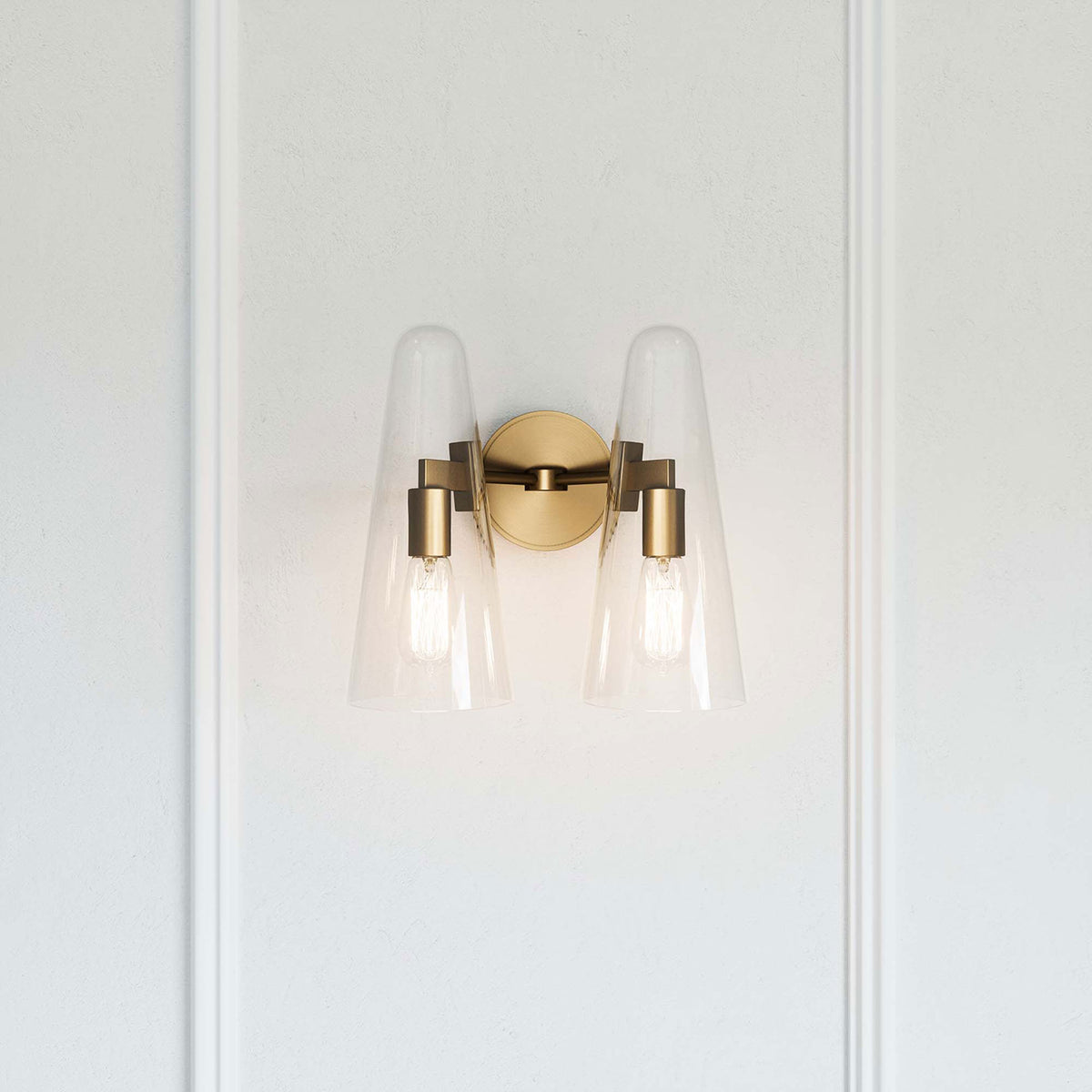 Beacon satin brass 2-light wall sconce casting a warm glow against a paneled white wall || EEI-5646-CLR-SBR || front_on_room
