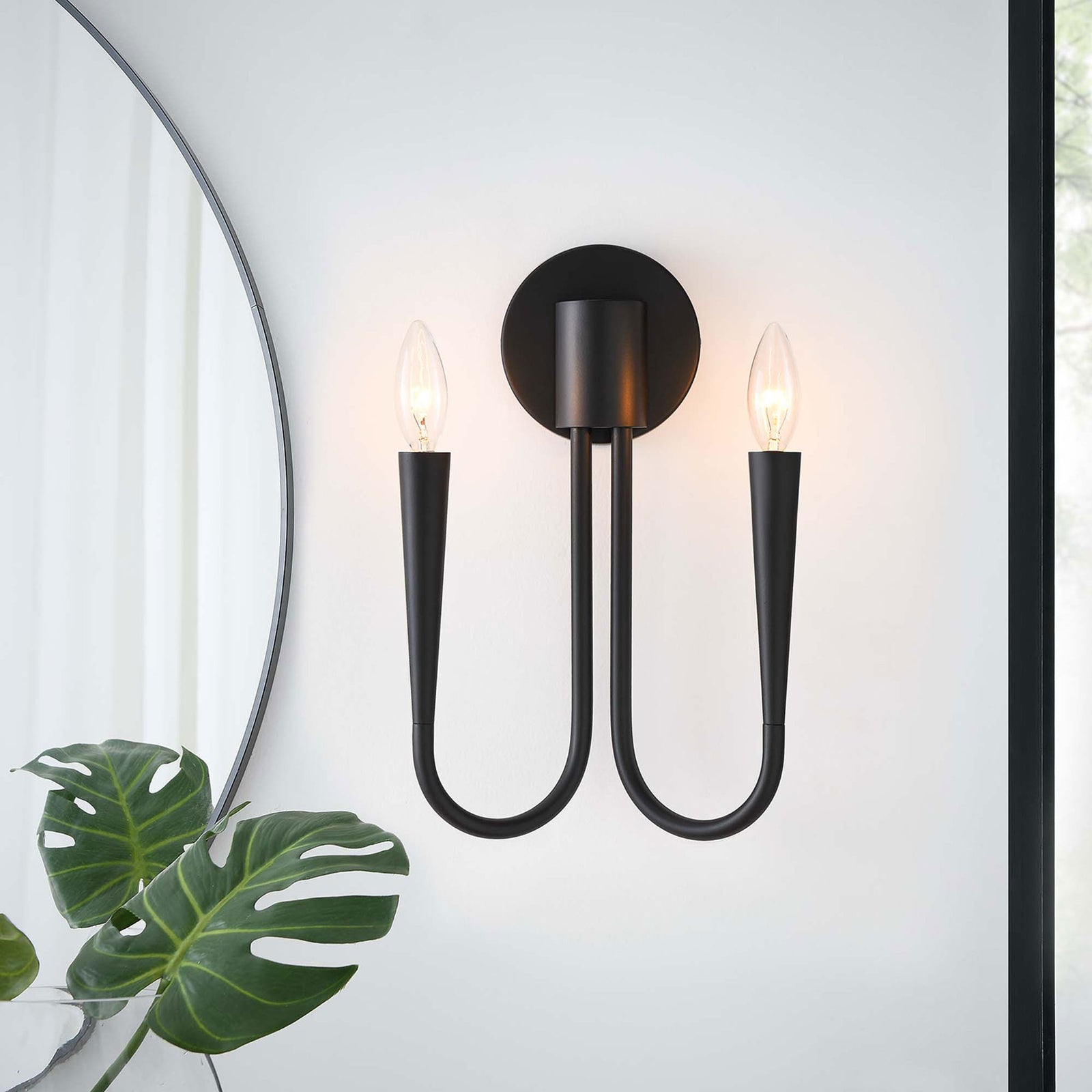 Penrose black 2-light wall sconce casts a warm glow beside a modern mirror, adding elegant ambiance to the room. || EEI-5666-BLK || front_angle_on_room