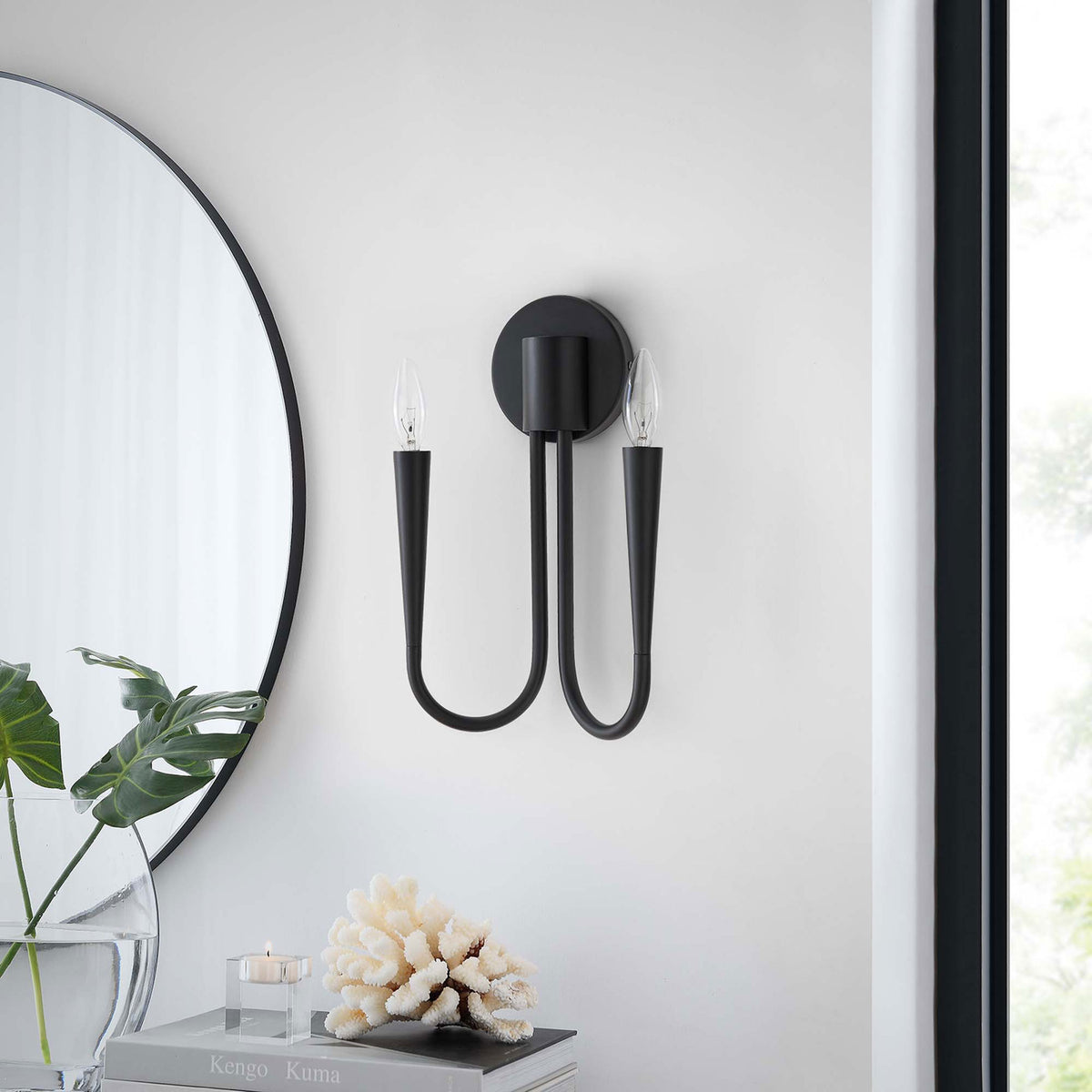 Unlit Penrose black wall sconce complements contemporary decor, pairing with greenery, books, and coral accents. || EEI-5666-BLK || front_room