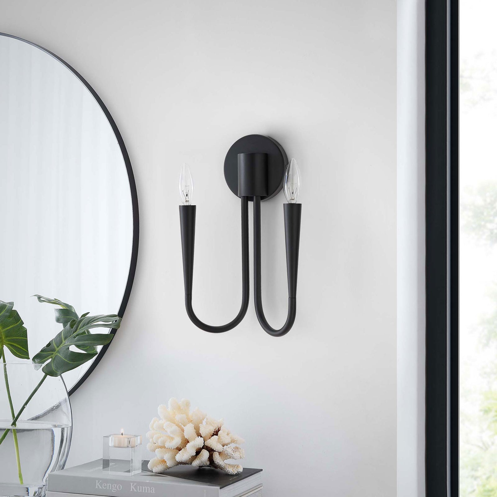 Unlit Penrose black wall sconce complements contemporary decor, pairing with greenery, books, and coral accents. || EEI-5666-BLK || front_room