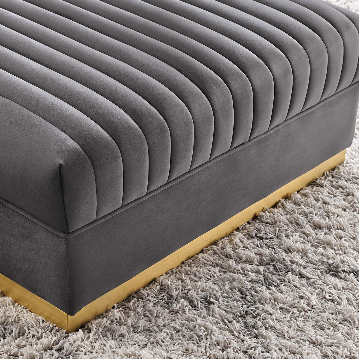 Gray channel tufted velvet ottoman resting on a plush shag rug, showcasing its lustrous gold base trim. || EEI-6036-GRY || detail_room