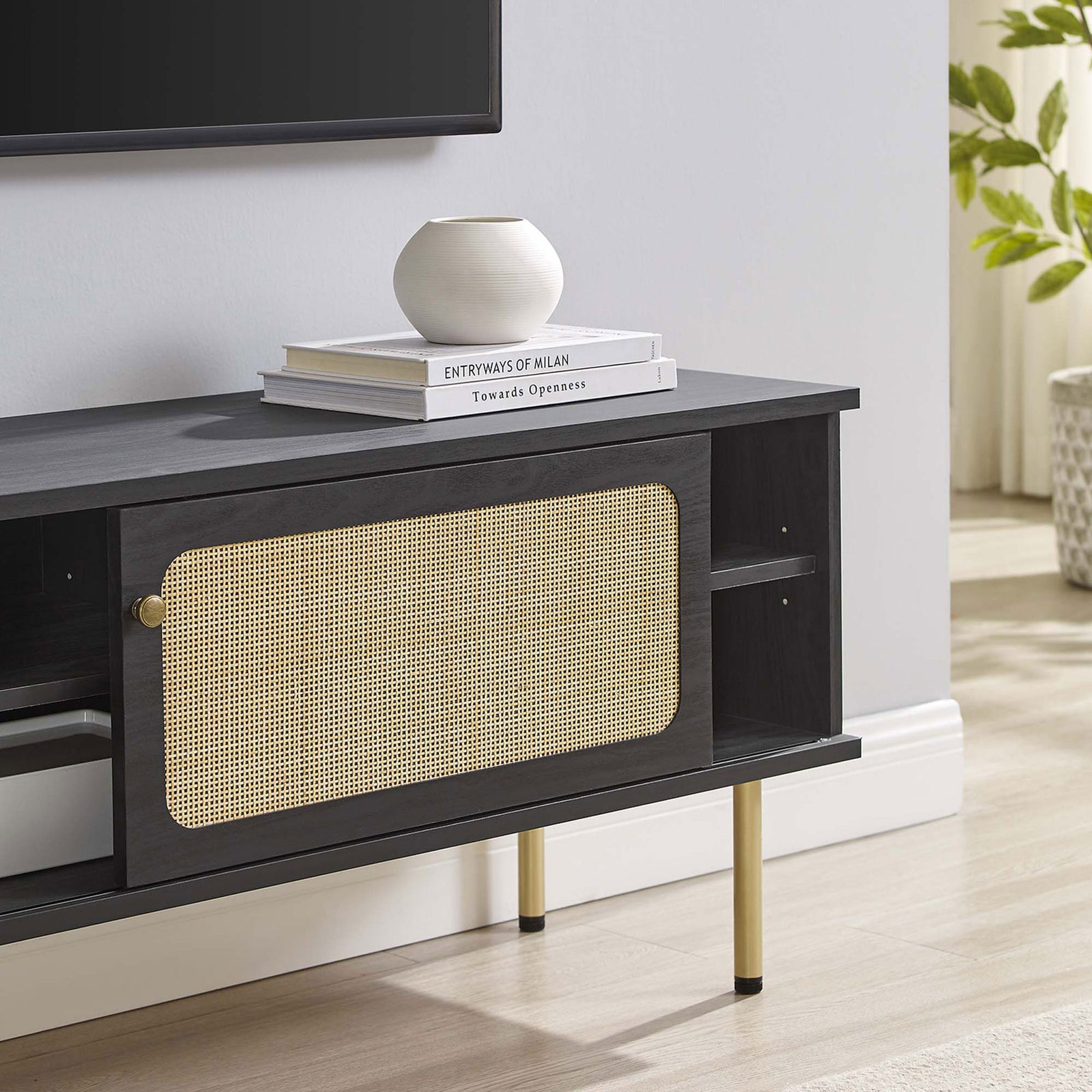 Black rattan TV stand styled under a wall-mounted TV with books and decor in a bright living room. || EEI-6044-BLK || storage_room
