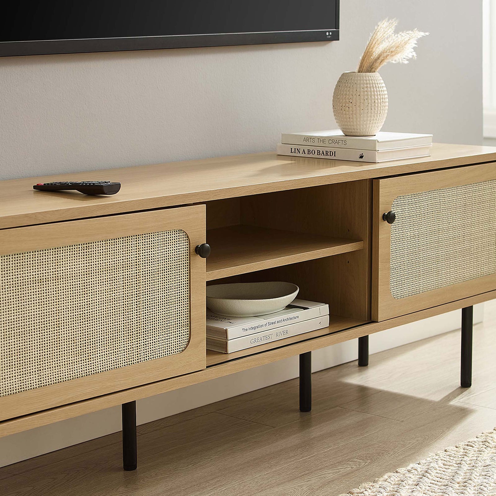 Oak rattan TV stand styled beneath a wall-mounted TV with books and decor in a bright living space. || EEI-6045-OAK || storage_room