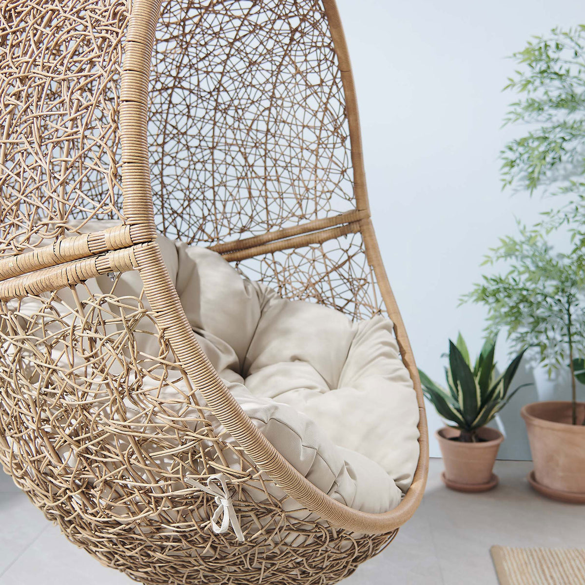 Beige cushioned swing chair basket brings cozy texture to an airy outdoor space with potted plants. || EEI-6262-CAP-BEI || detail_room