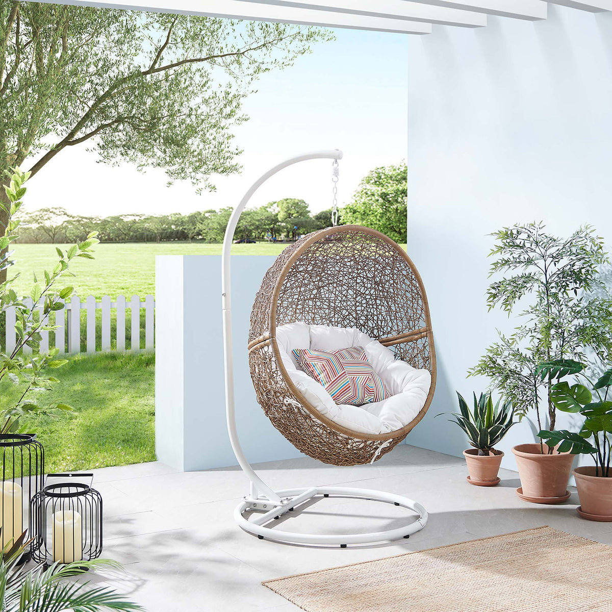 Cappuccino rattan swing chair with bright white cushion anchors a cheerful, greenery filled outdoor patio. || EEI-6262-CAP-WHI || front_angle_room