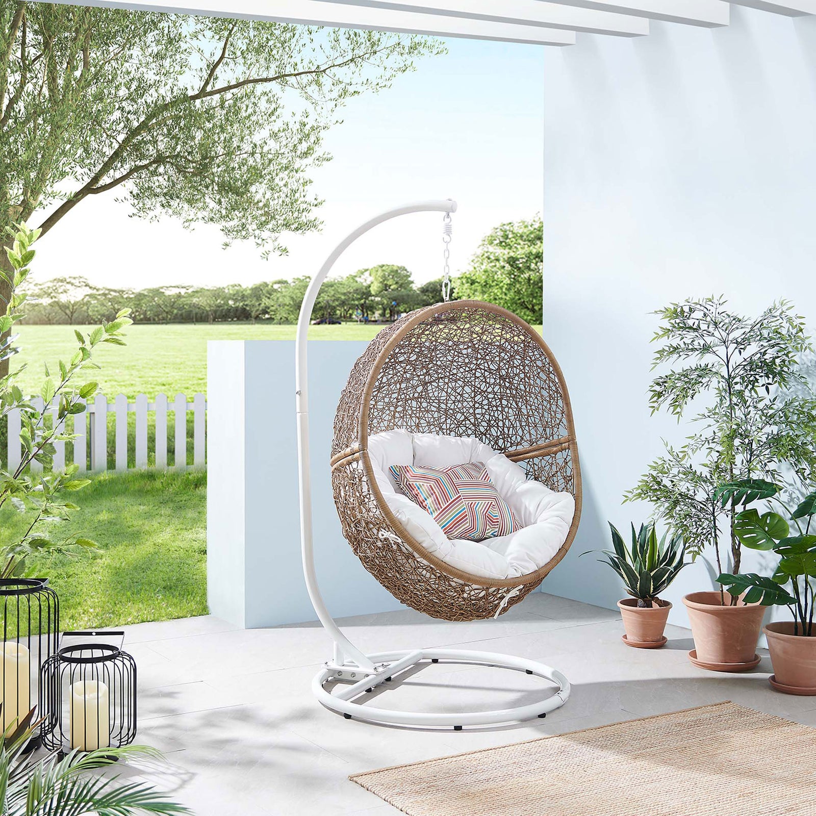 Cappuccino rattan swing chair with bright white cushion anchors a cheerful, greenery filled outdoor patio. || EEI-6262-CAP-WHI || front_angle_room