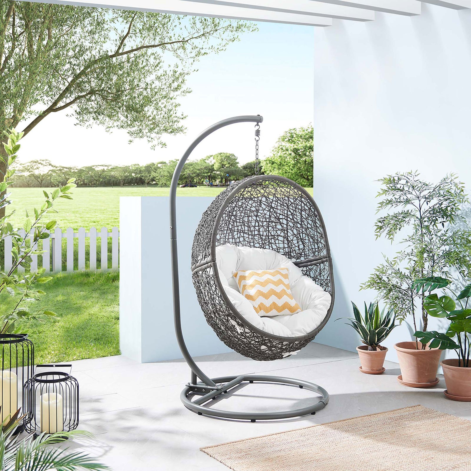 Gray rattan swing chair with white cushion creates a cozy focal point on a bright, open patio. || EEI-6262-GRY-WHI || front_angle_room
