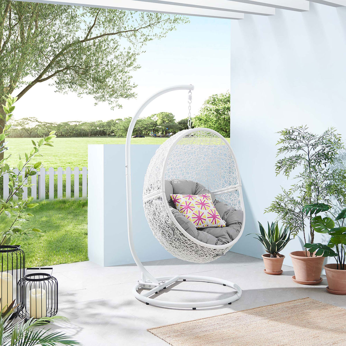 White rattan swing chair with gray cushion is styled on a fresh patio with greenery and soft light. || EEI-6262-WHI-GRY || front_angle_room