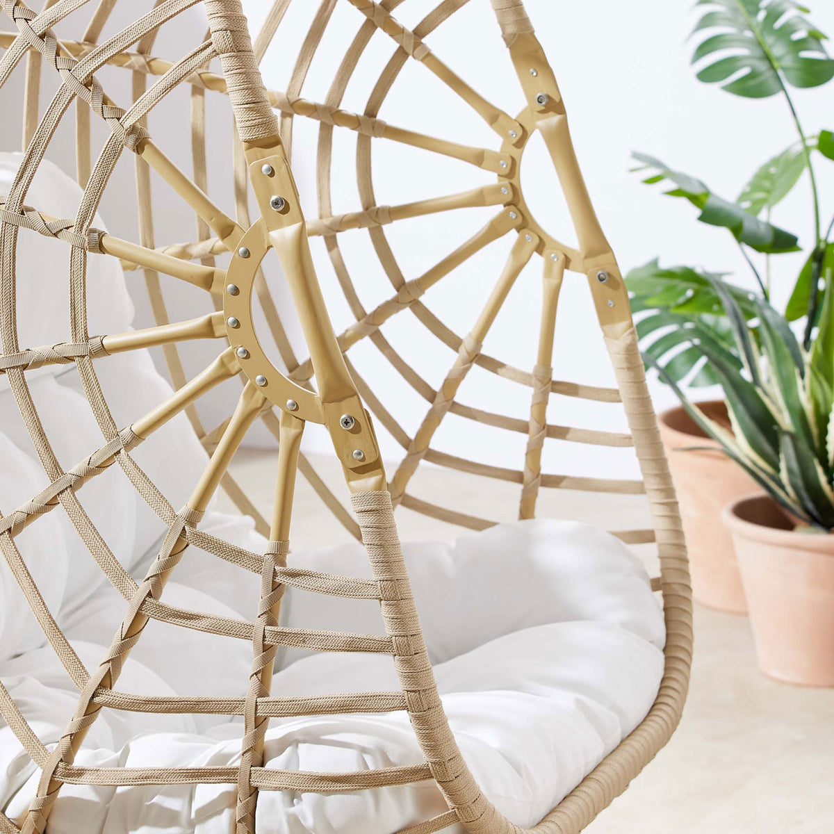 Rattan swing chair frame with neatly wrapped detailing and white cushion on a sunny patio with potted plants || EEI-6337-NAT-WHI || detail_room