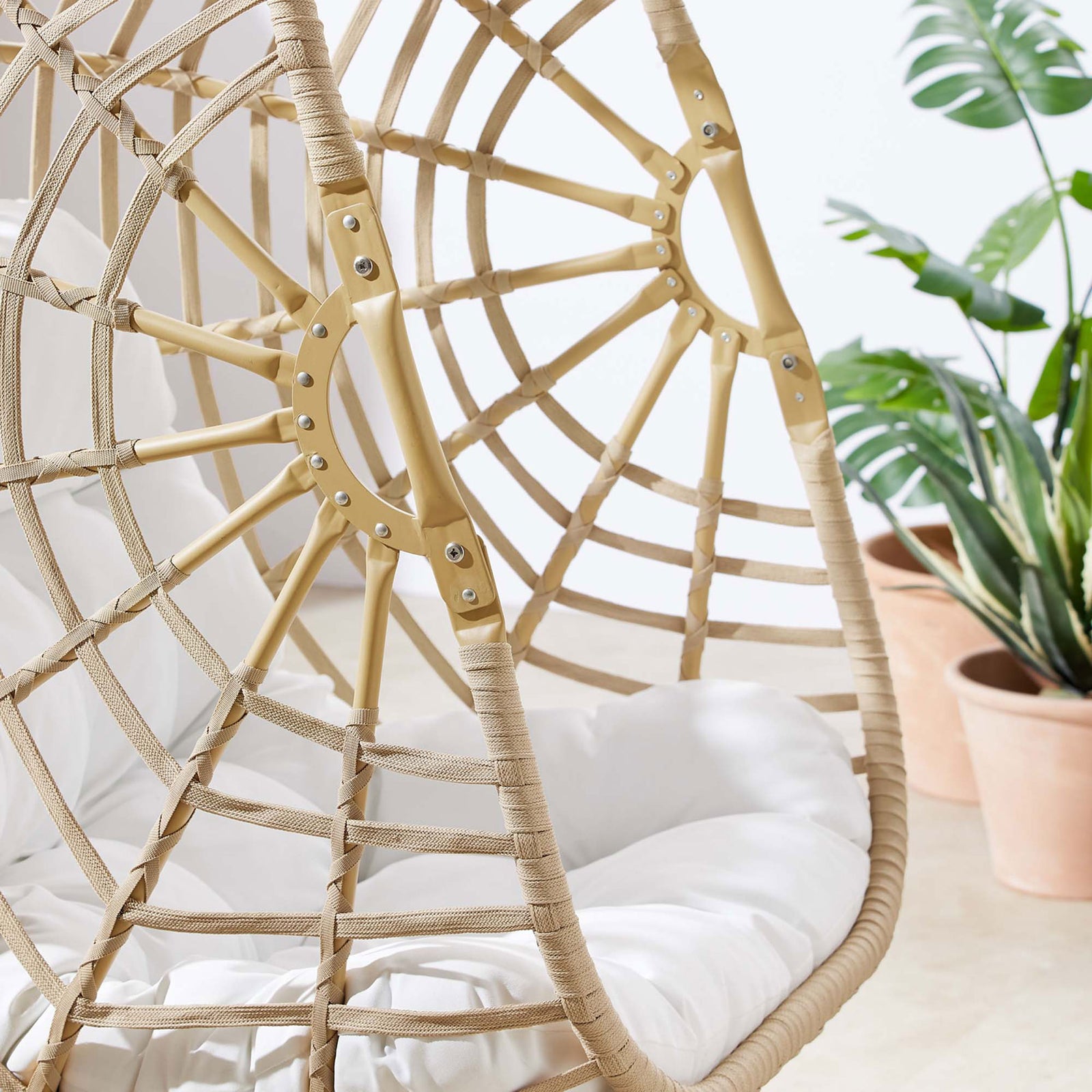 Rattan swing chair frame with neatly wrapped detailing and white cushion on a sunny patio with potted plants || EEI-6337-NAT-WHI || detail_room