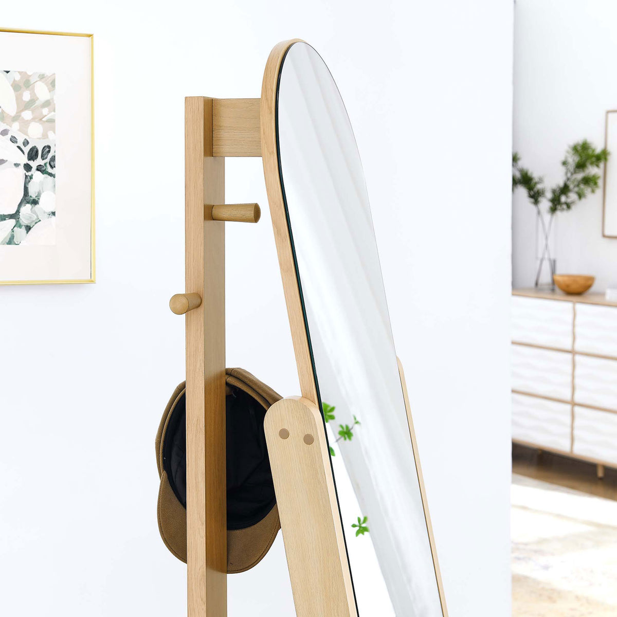 Oak standing mirror in a bright room with hat resting on side pegs and framed wall art || EEI-6346-OAK || detail_room