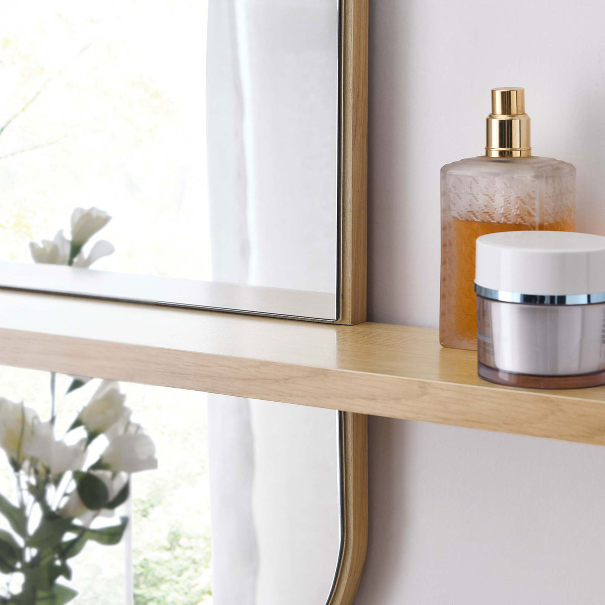 Oak mirror shelf holding skincare bottles beside soft florals and window light. || EEI-6349-OAK || storage_room