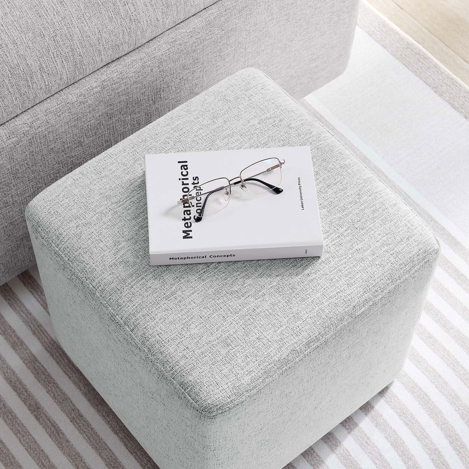 Ottoman used as a compact side table, topped with a book and glasses in a modern living space || EEI-6636-HLG || top_angle_room
