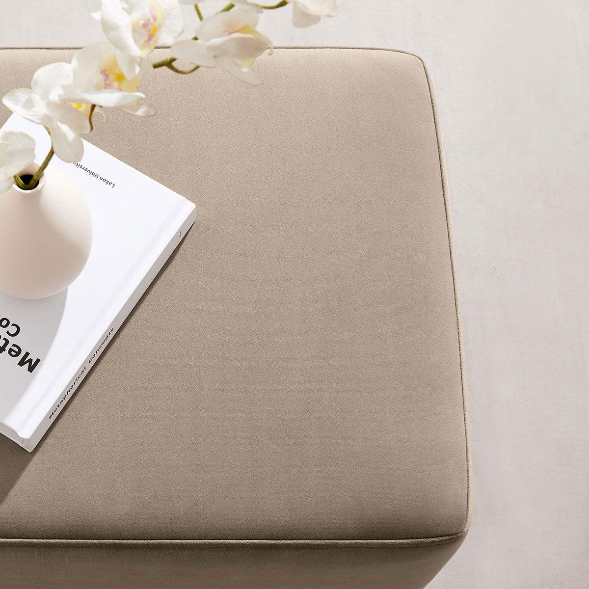 Taupe velvet ottoman styled as a chic side table with a vase and book in a bright, airy room. || EEI-6640-TAU-NAT || detail_room