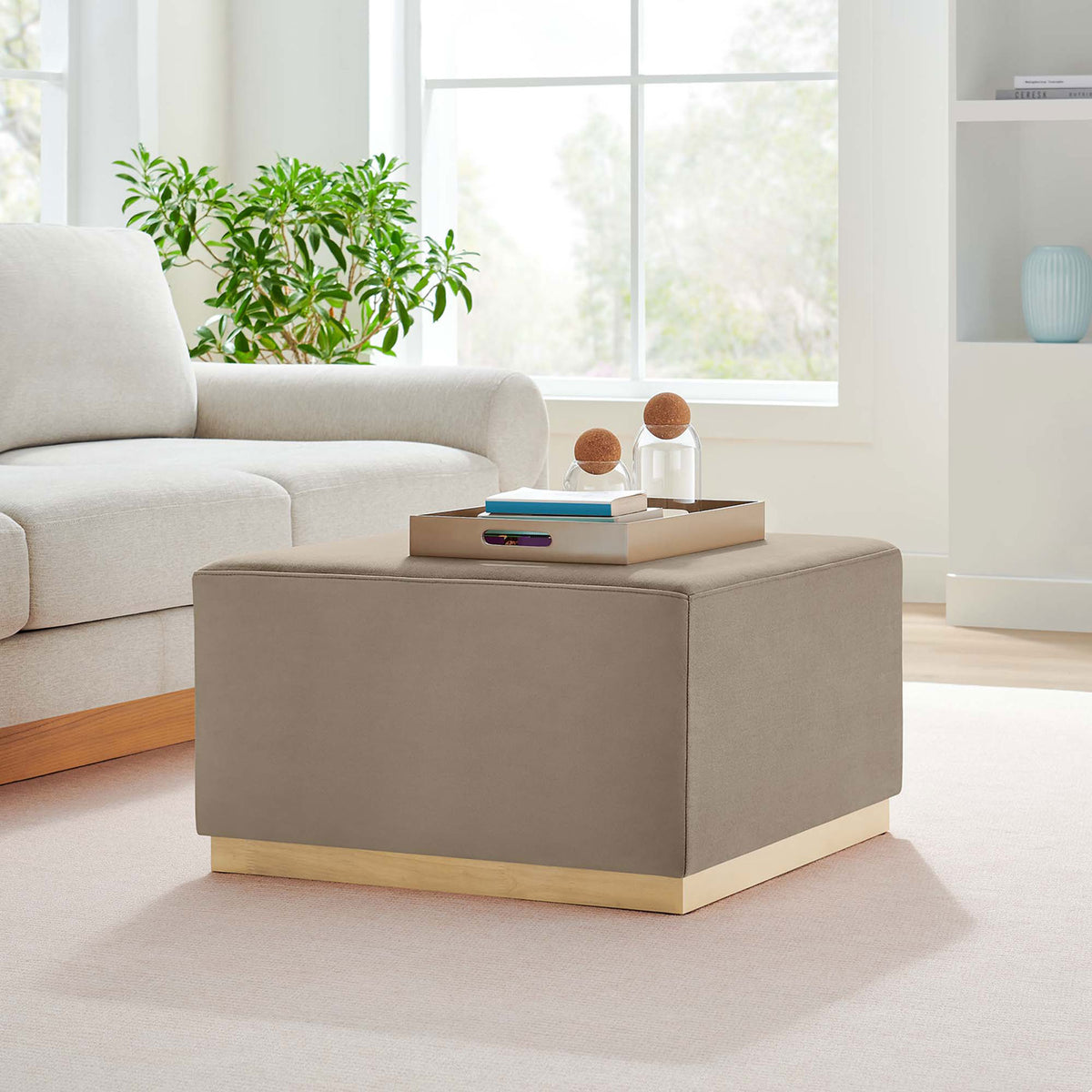 Taupe velvet square ottoman on wood base styled as a coffee table in a bright modern living room || EEI-6643-TAU-NAT || front_angle_room
