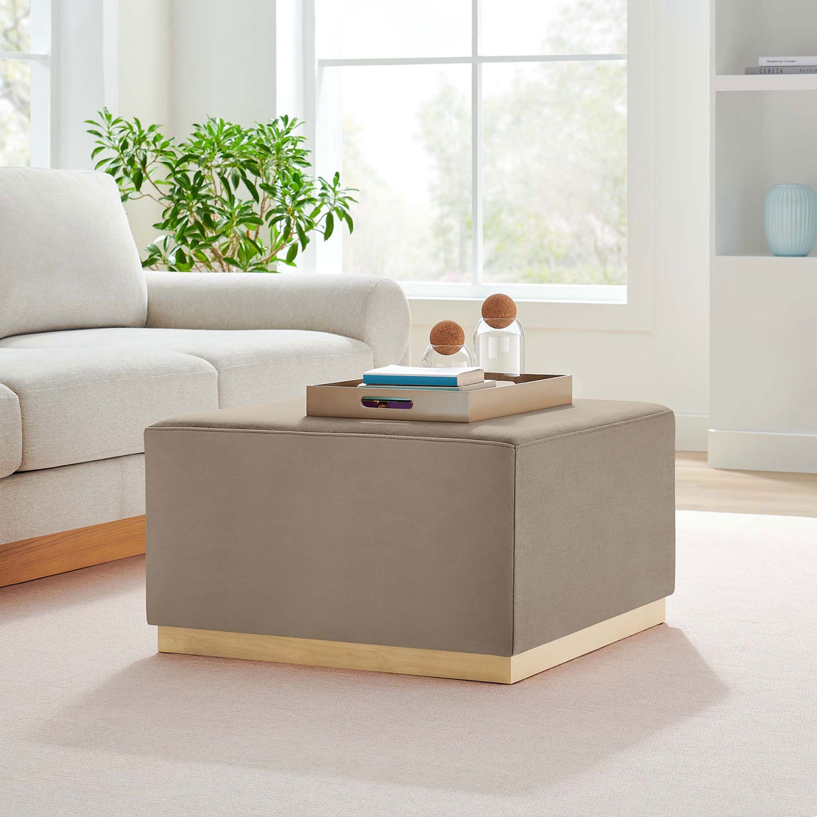 Taupe velvet square ottoman on wood base styled as a coffee table in a bright modern living room || EEI-6643-TAU-NAT || front_angle_room