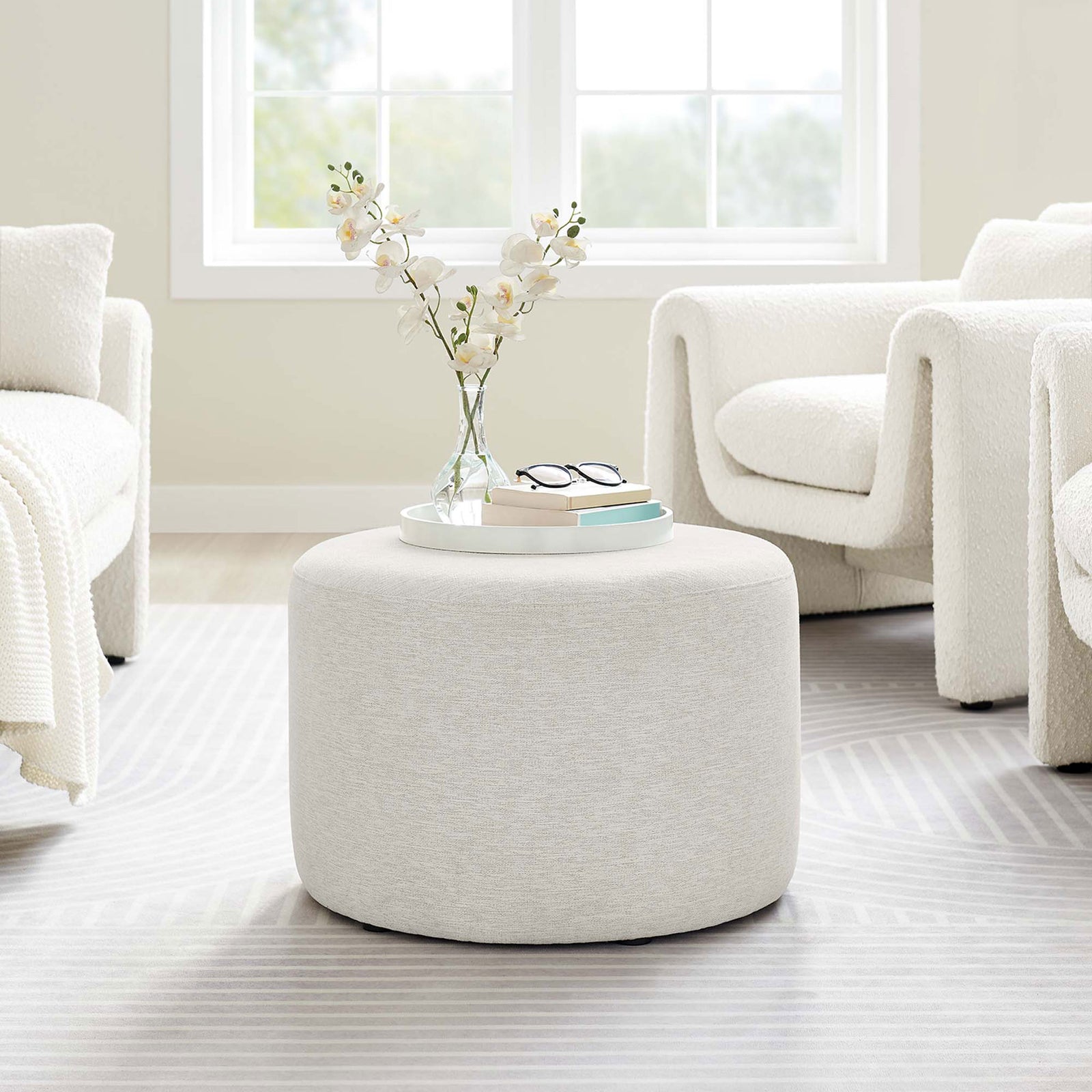 Ivory round ottoman used as a coffee table in a bright modern living room with curved seating || EEI-6645-HEI || front_angle_room