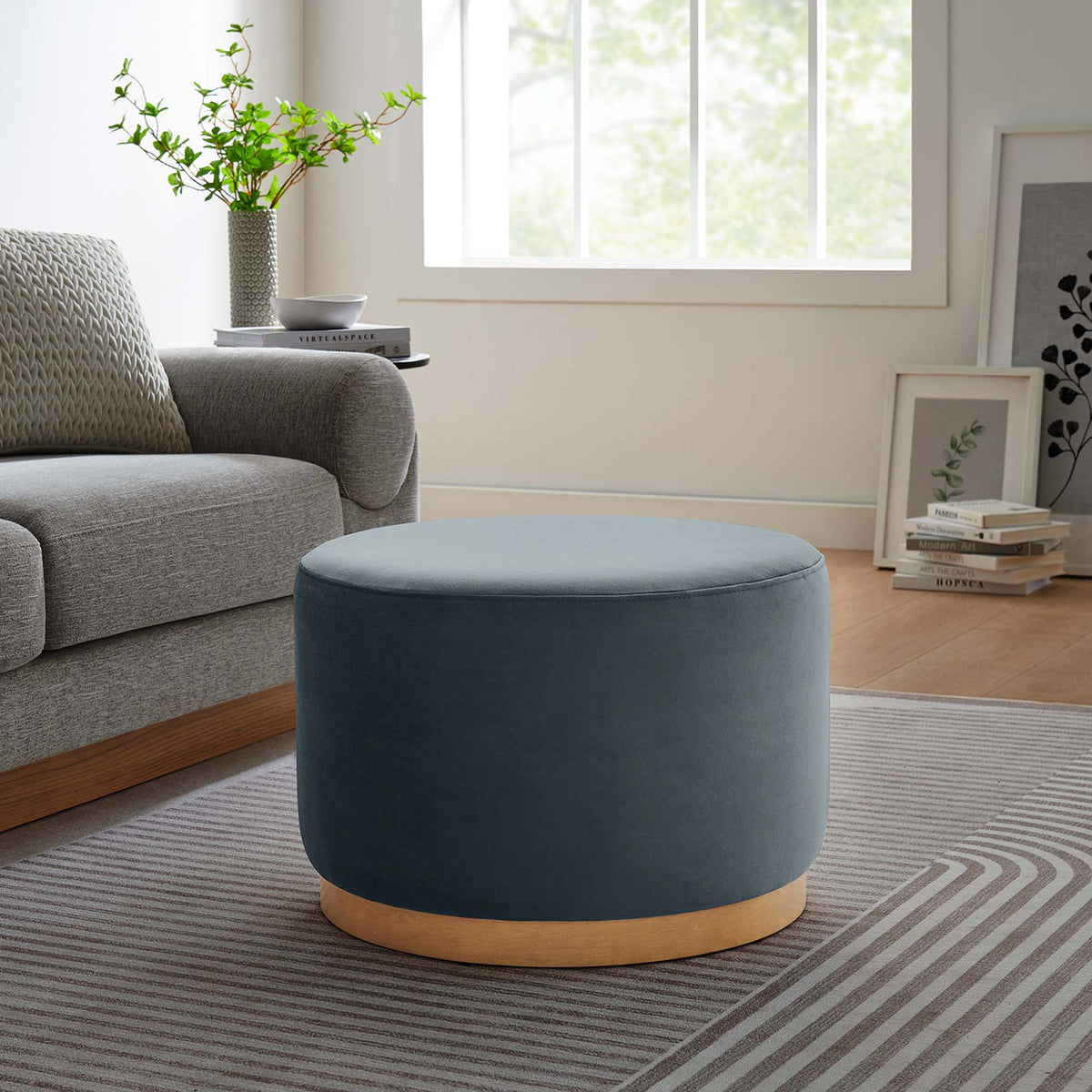 Round blue velvet ottoman with warm wood base complementing a contemporary living room with neutral decor || EEI-6646-CAB-NAT || front_angle_room