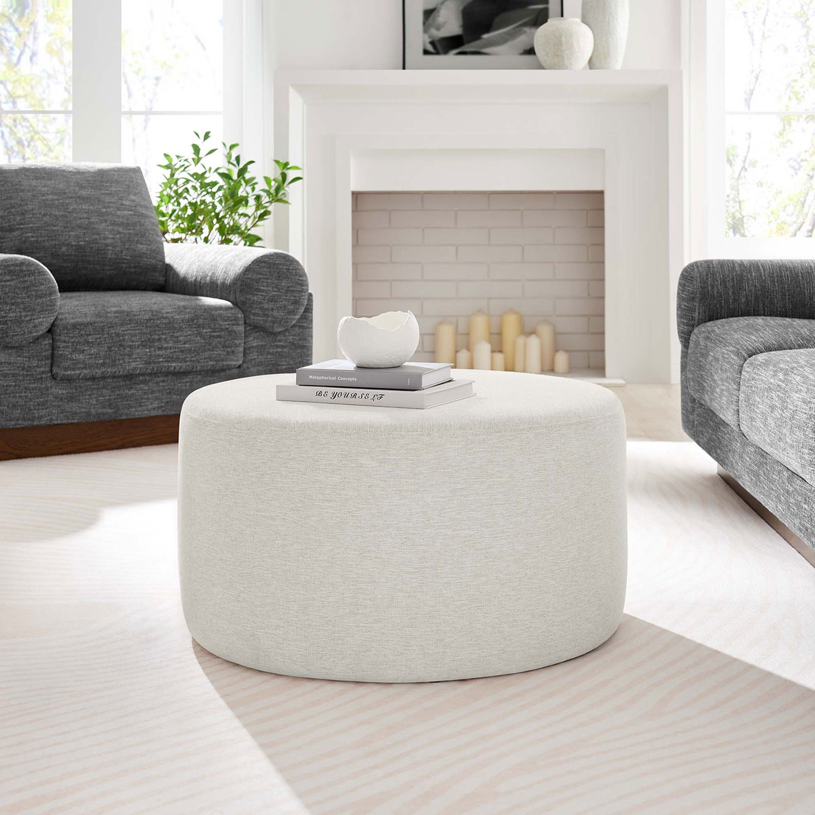Ivory round ottoman styled as a coffee table in a bright living room with gray sofas and a white fireplace. || EEI-6647-HEI || front_angle_room