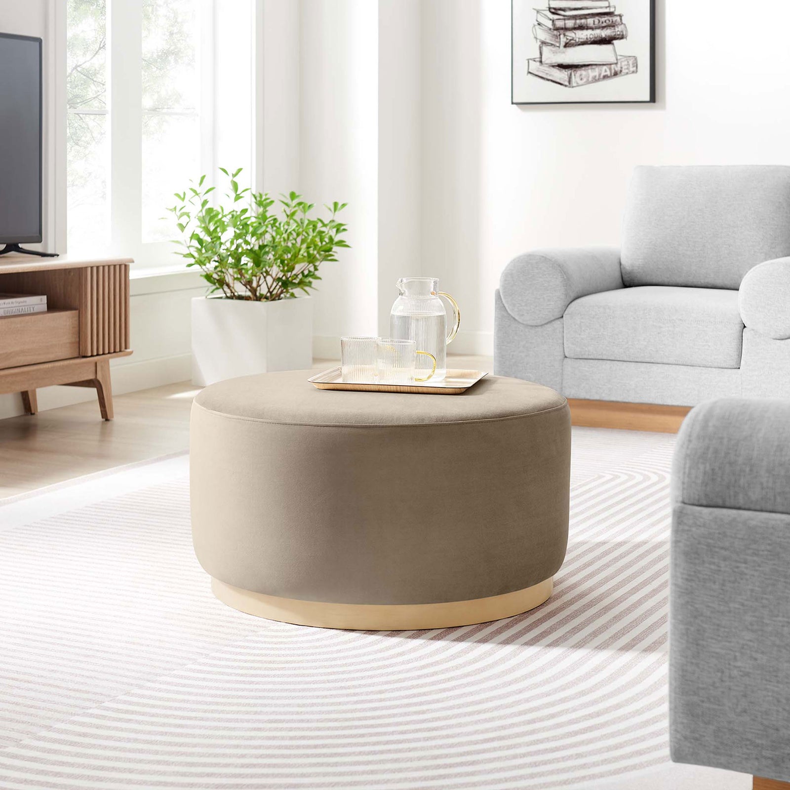 Taupe round velvet ottoman centered in a bright modern living room with neutral sofa and light wood accents || EEI-6648-TAU-NAT || front_room