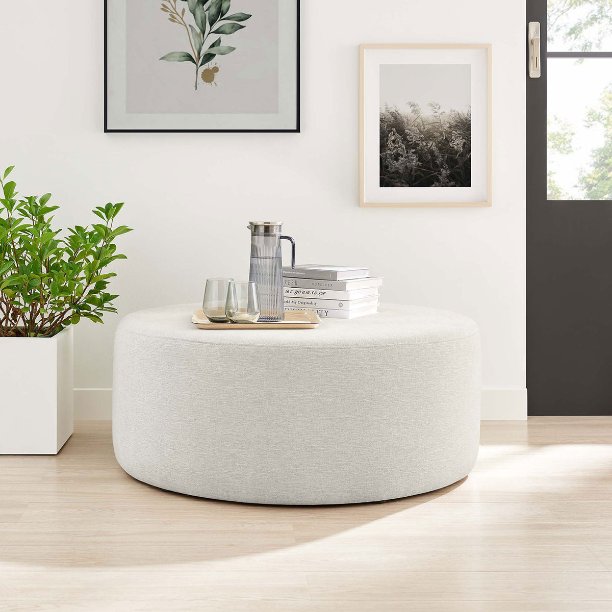 Generous ivory round ottoman used as a coffee table in a bright modern room with art and greenery. || EEI-6649-HEI || front_angle_room