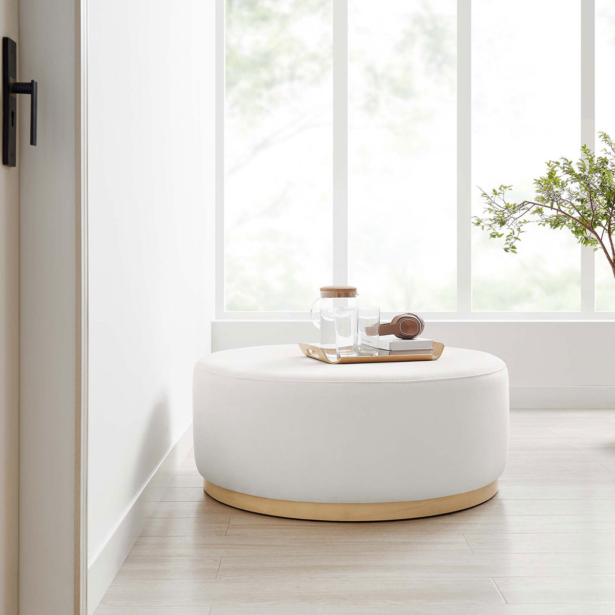 Tilden 38 inch round velvet ottoman used as a casual table in a bright minimalist room with greenery || EEI-6650-ALA-NAT || front_angle_room