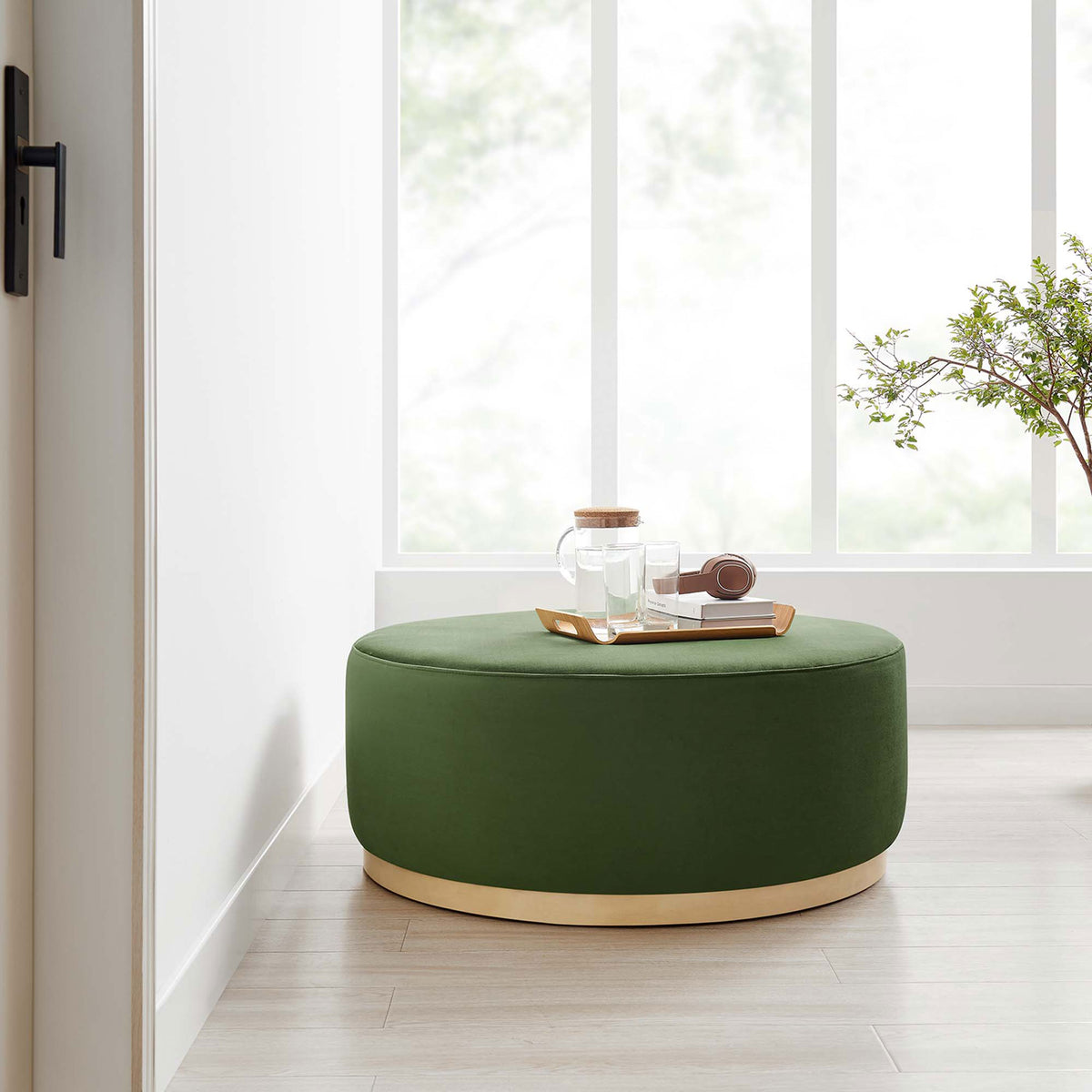 Generous moss green velvet round ottoman with wood base serving as a coffee table in a bright airy room. || EEI-6650-MOG-NAT || front_angle_room