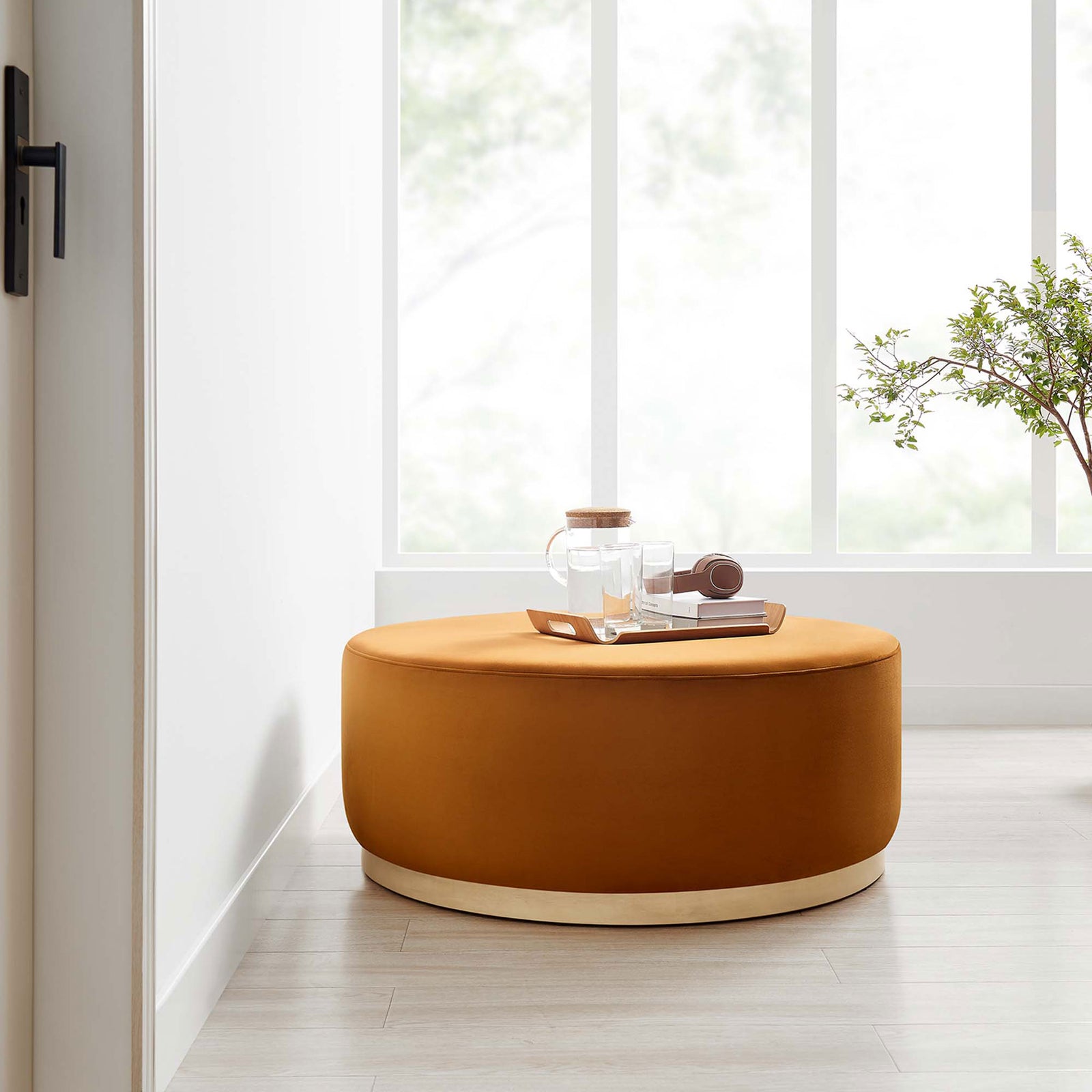 Large rust velvet round ottoman with wood base placed in an airy light filled room. || EEI-6650-RUS-NAT || front_angle_room