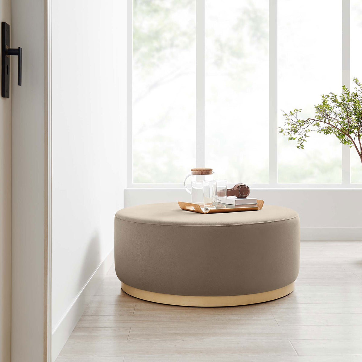 Taupe round ottoman styled in a bright windowed room with tray and decor on a natural wood floor || EEI-6650-TAU-NAT || front_room
