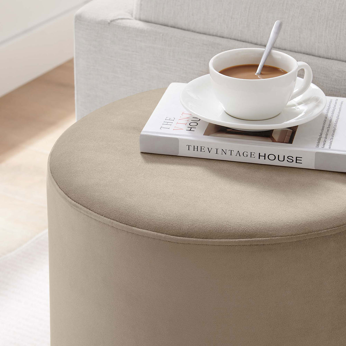 Round taupe ottoman used as a chic side table beside a sofa with coffee and a book || EEI-6652-TAU-NAT || front_angle_room